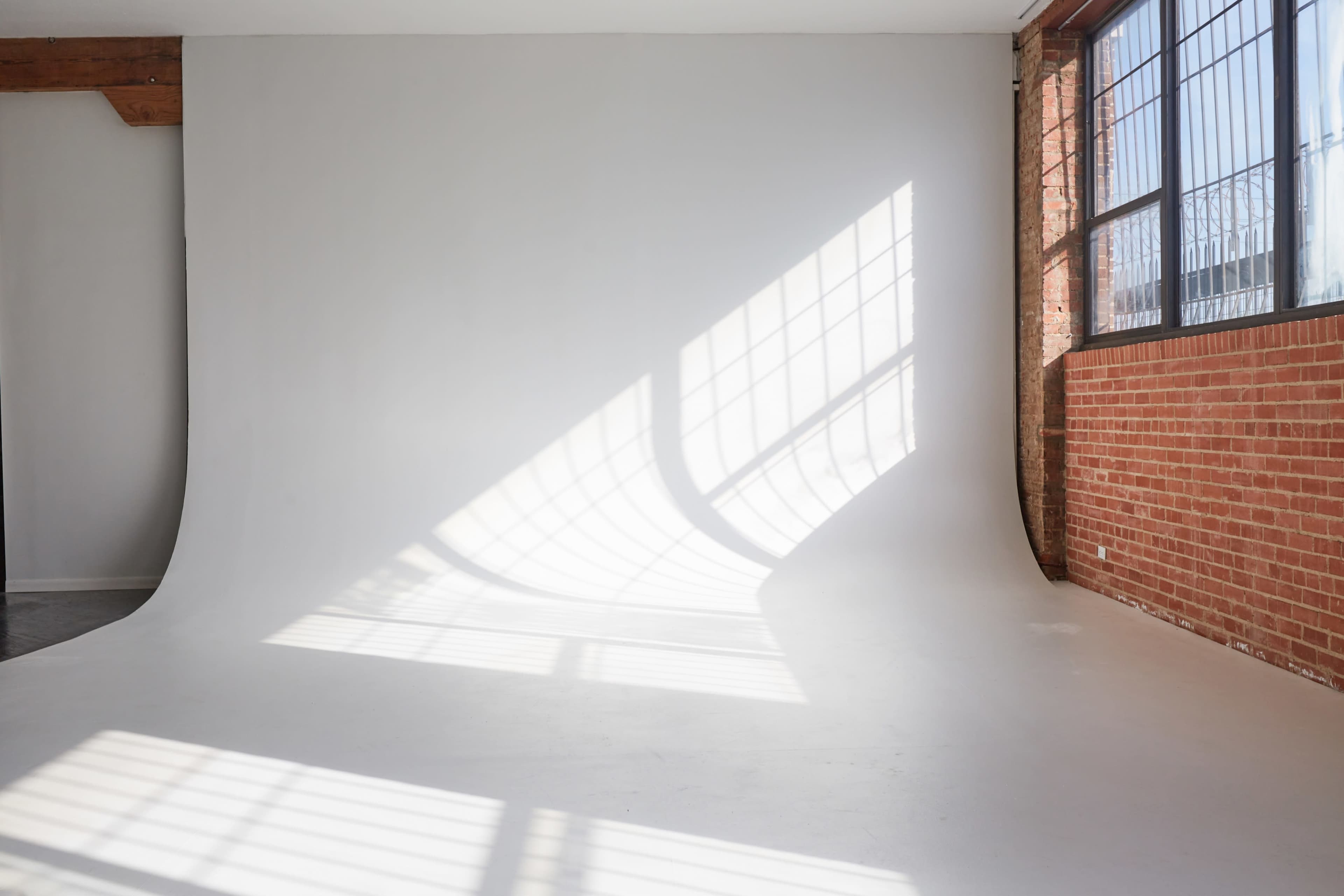 A bright, minimalist studio space with a white backdrop and sunlight casting a pattern on the floor from a window with grid-style panes.