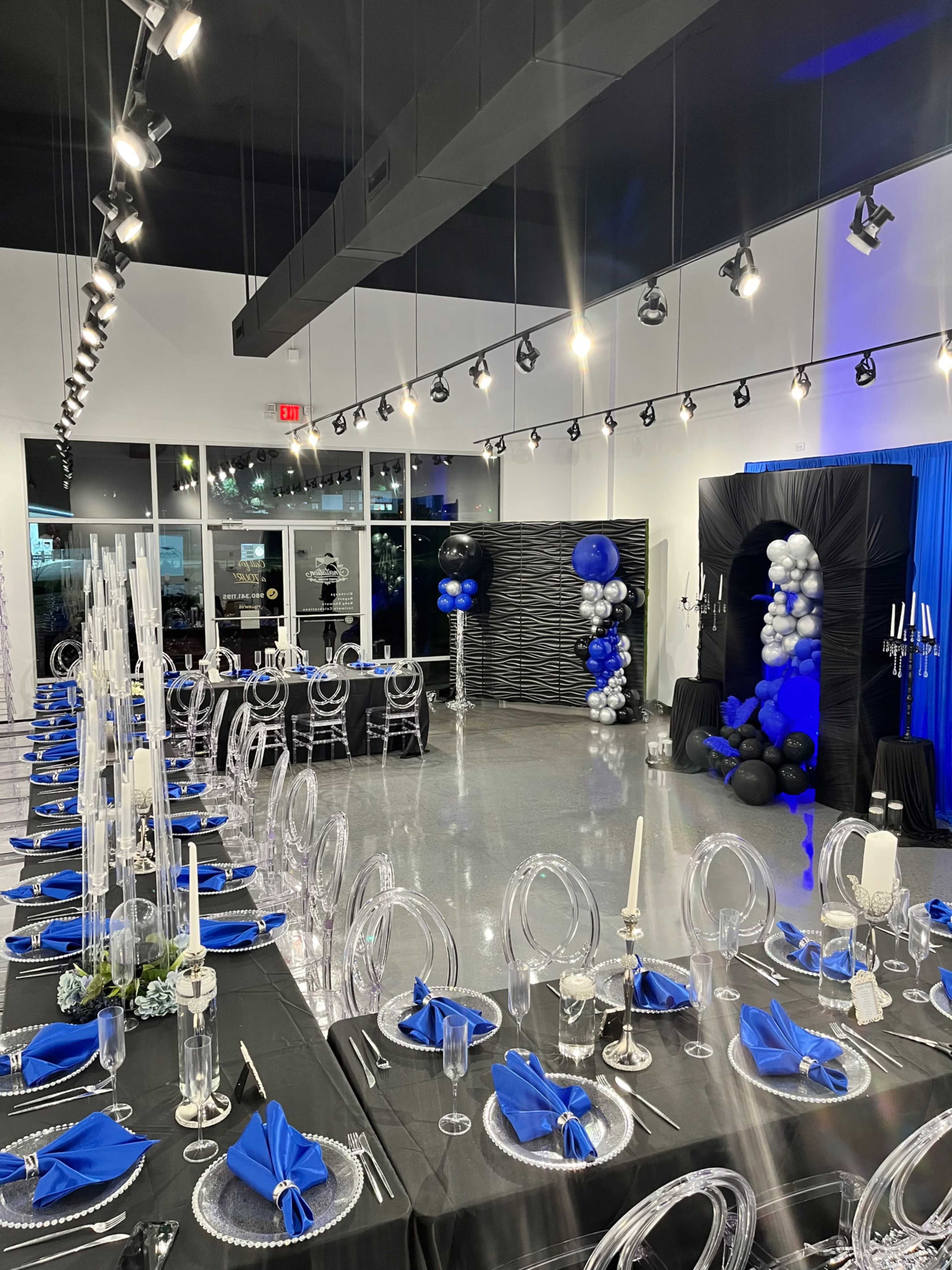 The image shows a banquet hall set up for a formal event, featuring long, elegantly arranged tables with black tablecloths, blue napkins, and clear dining chairs, alongside decorative balloons and a backdrop.