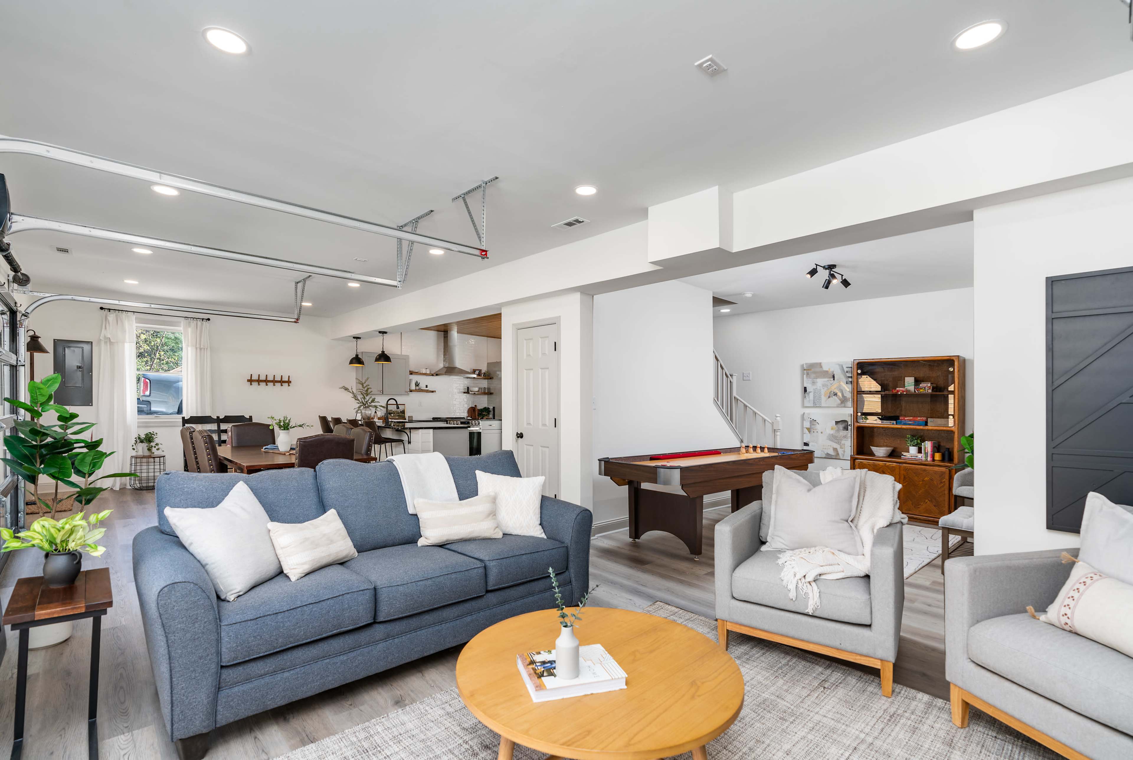A modern living room with a gray sofa, two armchairs, a round wooden coffee table, and a view of a game area with a pool table and a kitchen in the background.