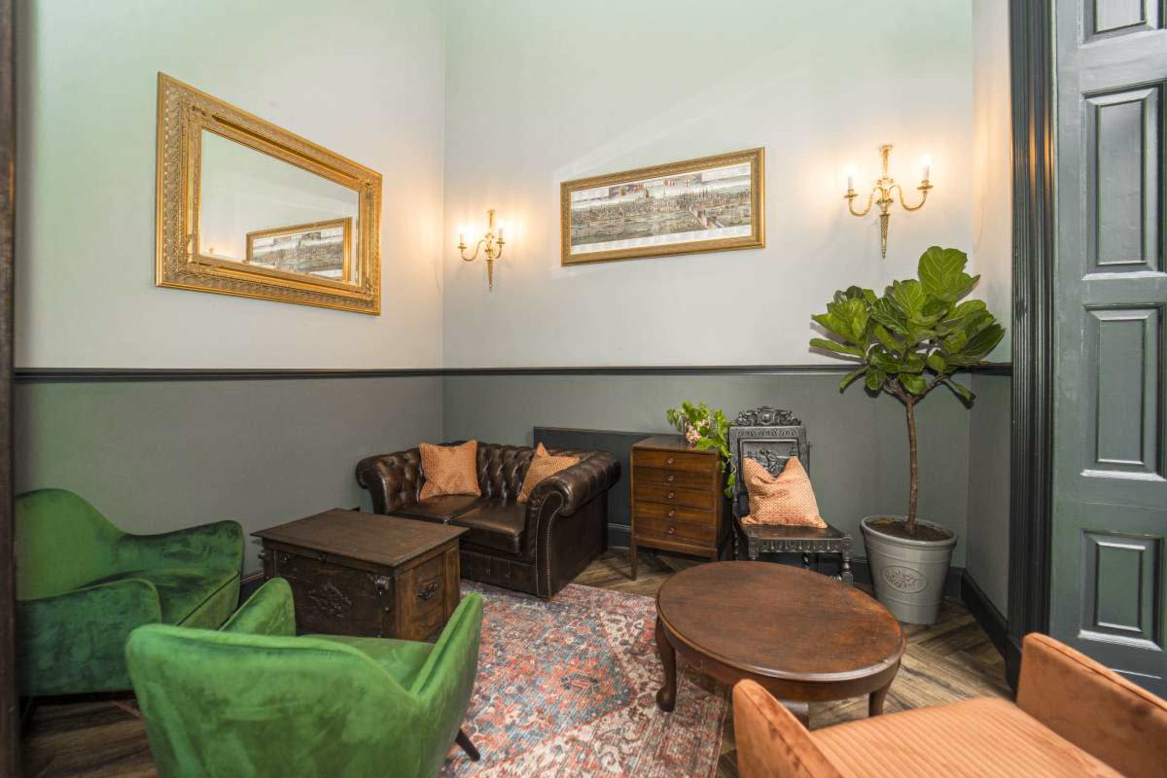 The image depicts a cozy sitting area with a dark leather couch, green armchairs, a wooden coffee table, and a decorative plant beside a vintage-style mirror and artwork on the wall.