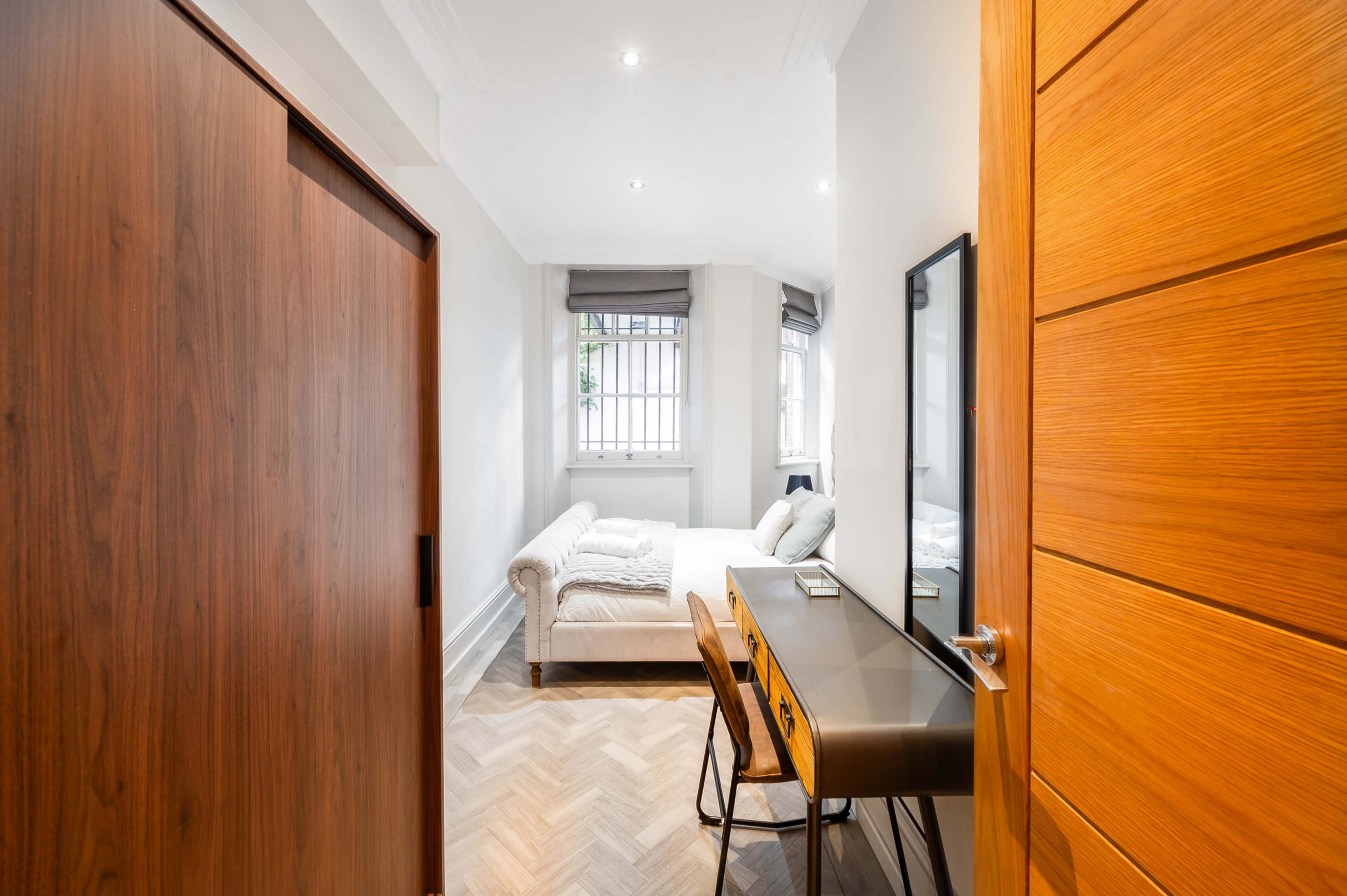 A small room features a bed with white linens, a wooden writing desk with chairs, and a large window allowing natural light.