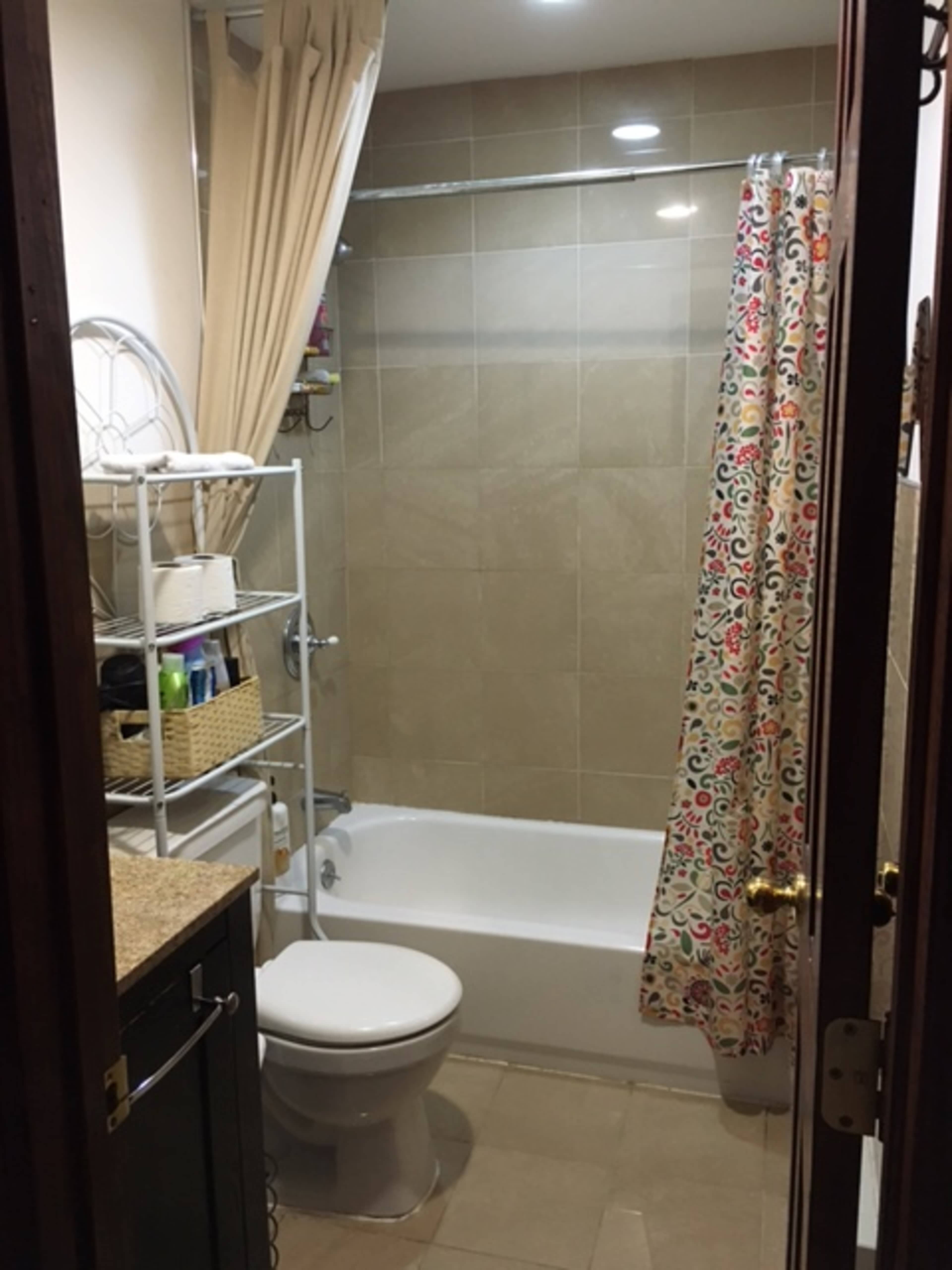 A small bathroom featuring a bathtub with a shower curtain, a toilet, and a shelving unit with toiletries.