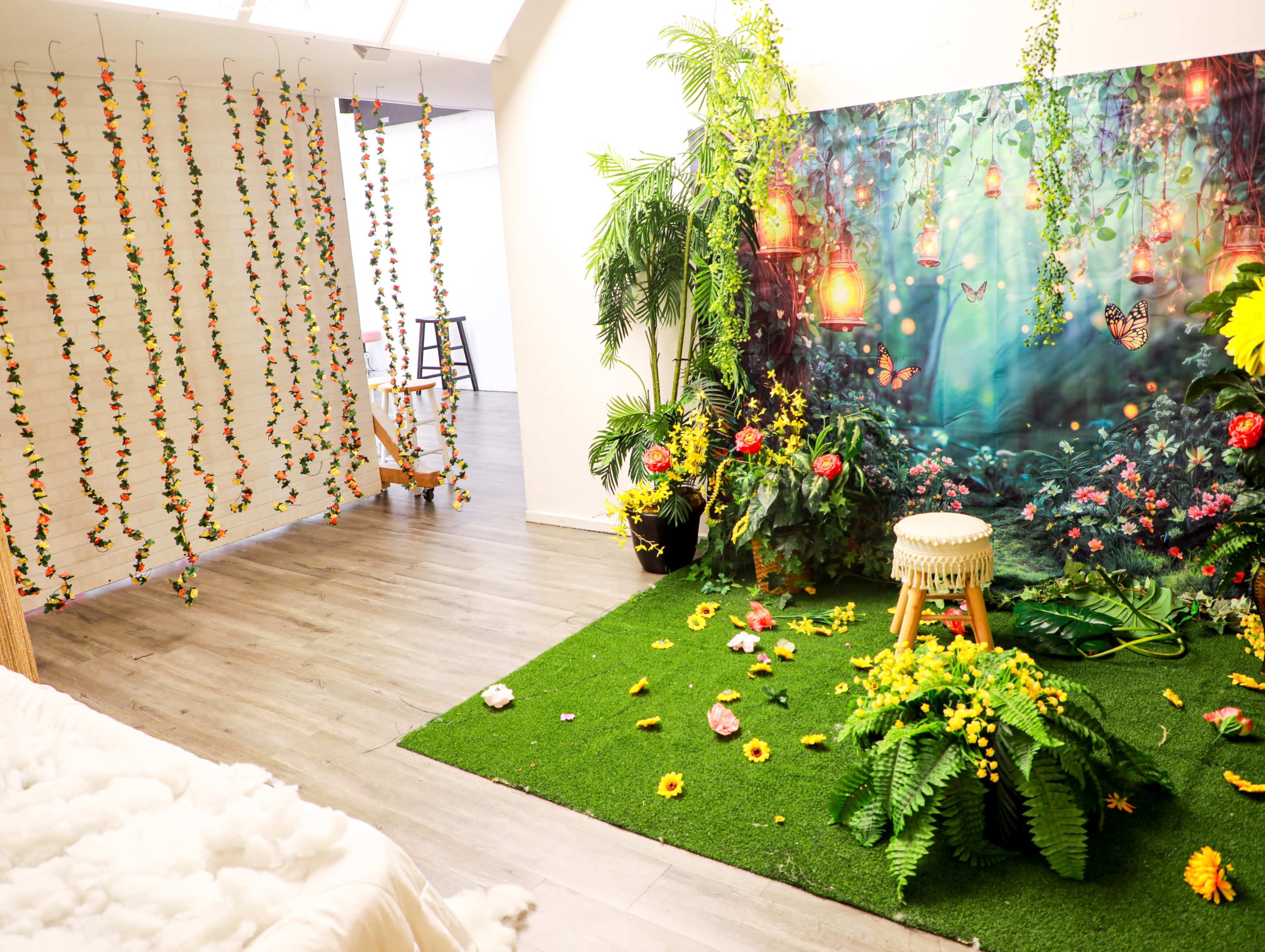 The image features a vibrant photo studio with lush green artificial grass, a floral backdrop with lanterns, and decorative plants arranged alongside a wooden stool and scattered flowers.