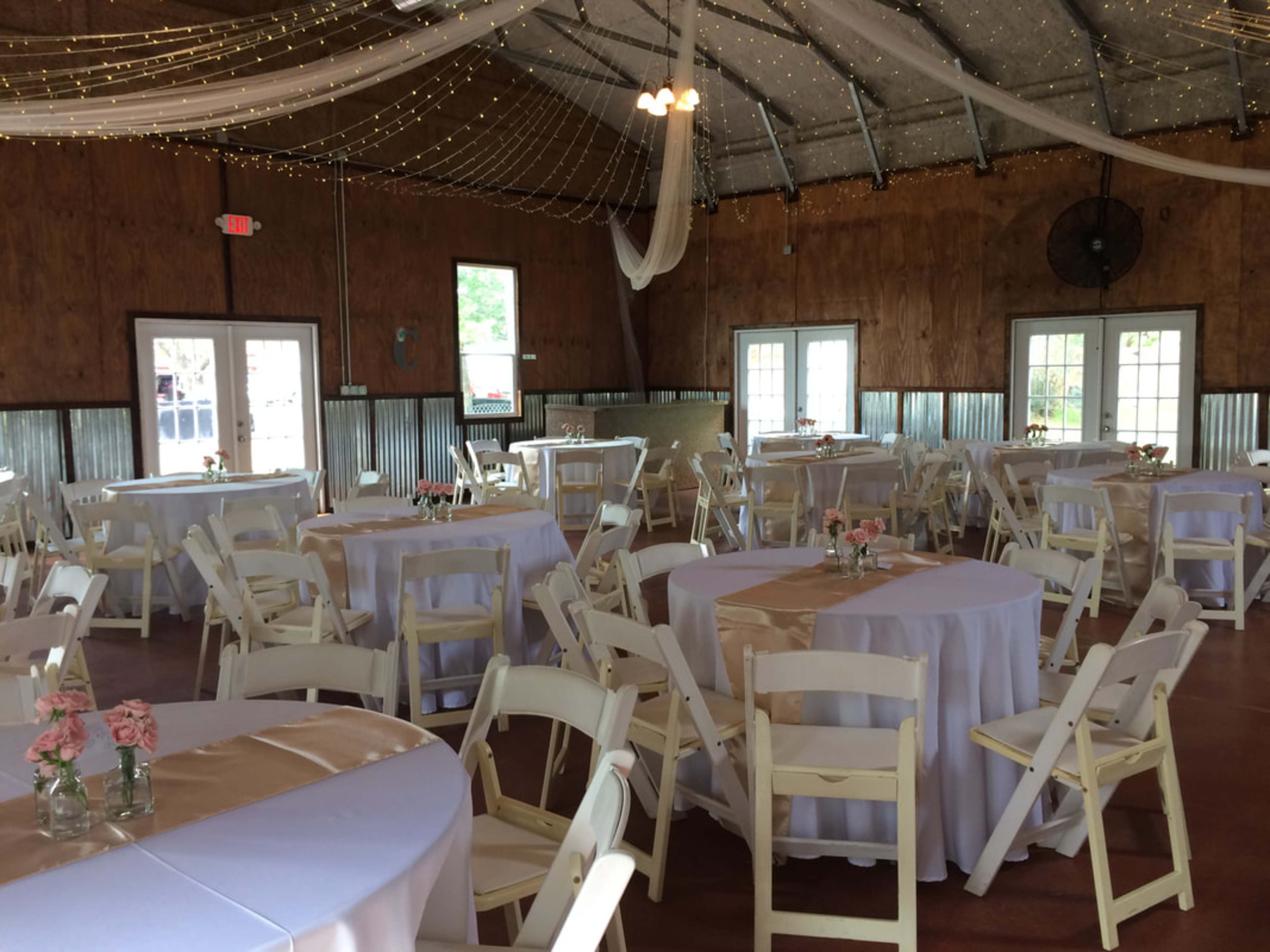 The image shows a spacious event venue with round tables covered in white tablecloths and pink decorative elements, surrounded by a wooden interior and string lights overhead.