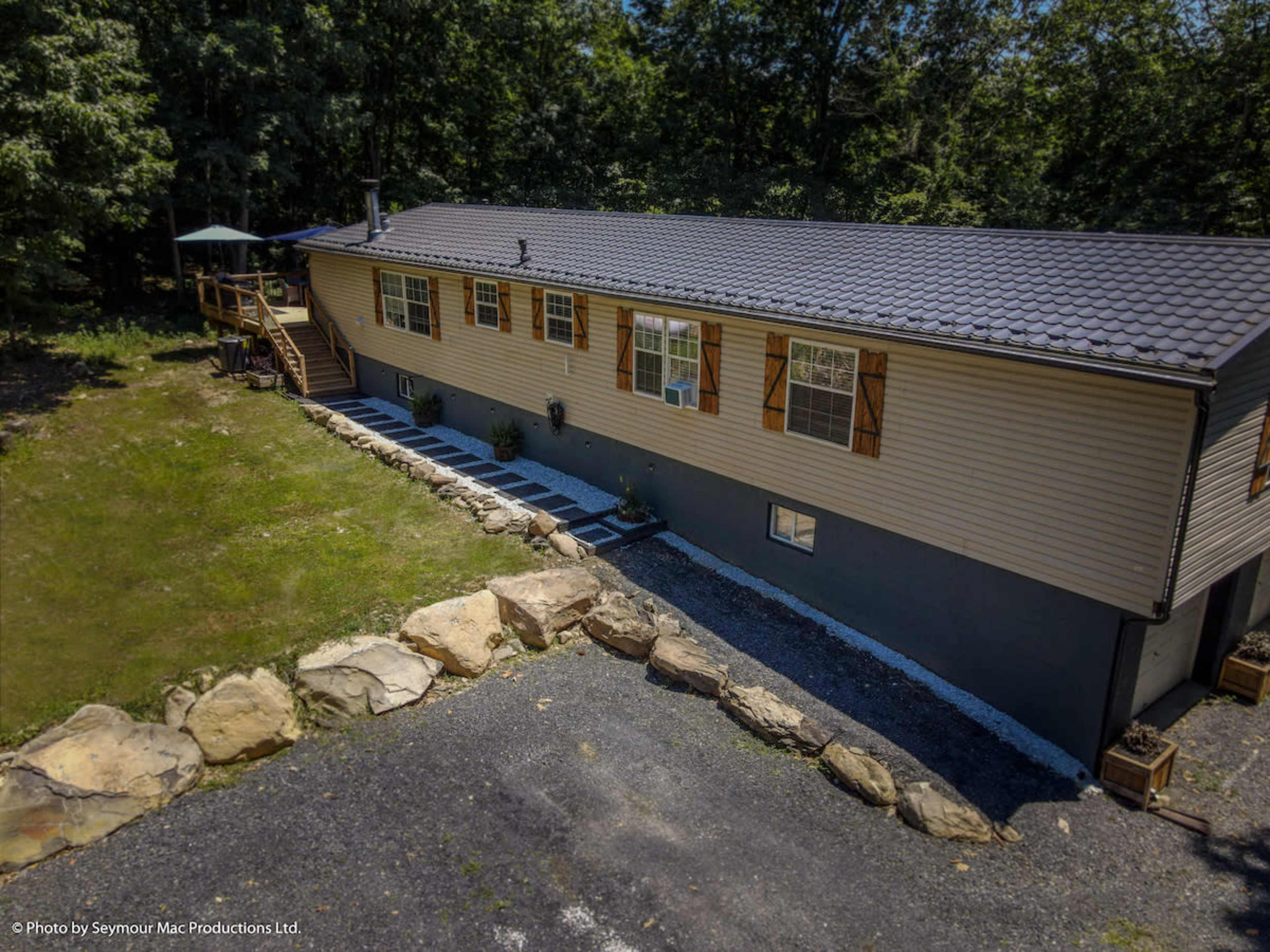 A single-story house with gray siding and a shale stone patio is surrounded by trees and a gravel driveway.