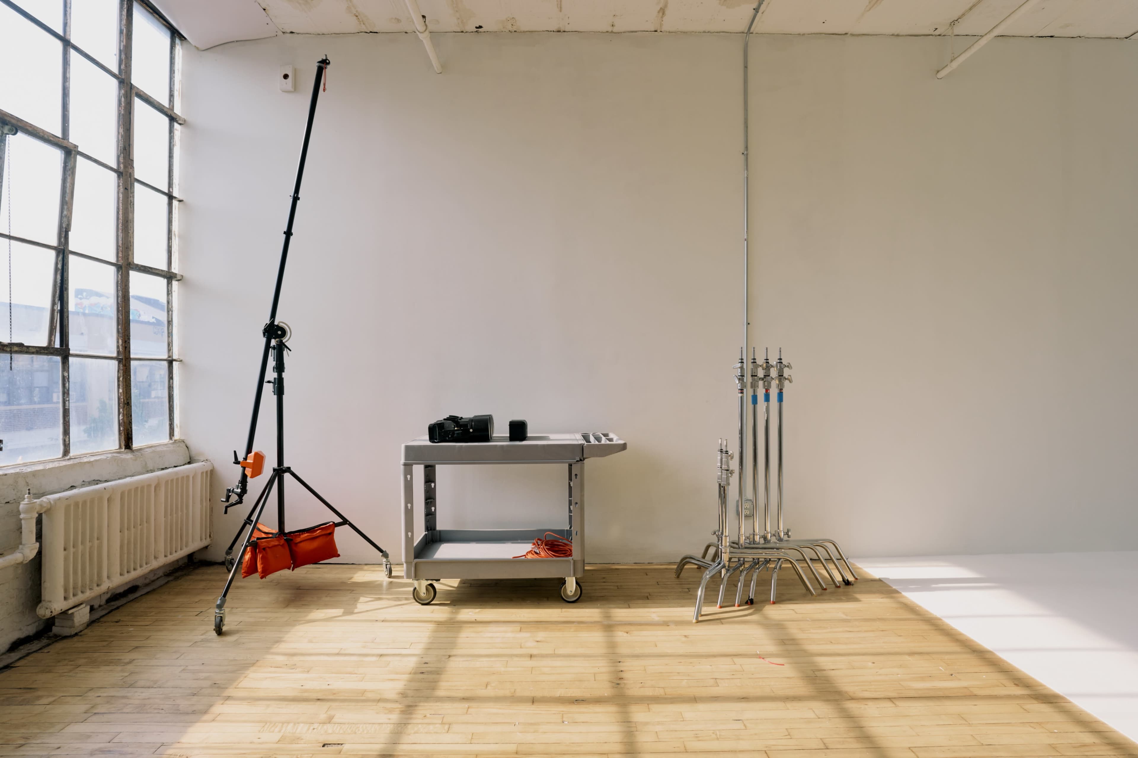 The image shows a minimalist indoor space featuring a table on wheels, a camera, and several lighting stands along with a backdrop support system.