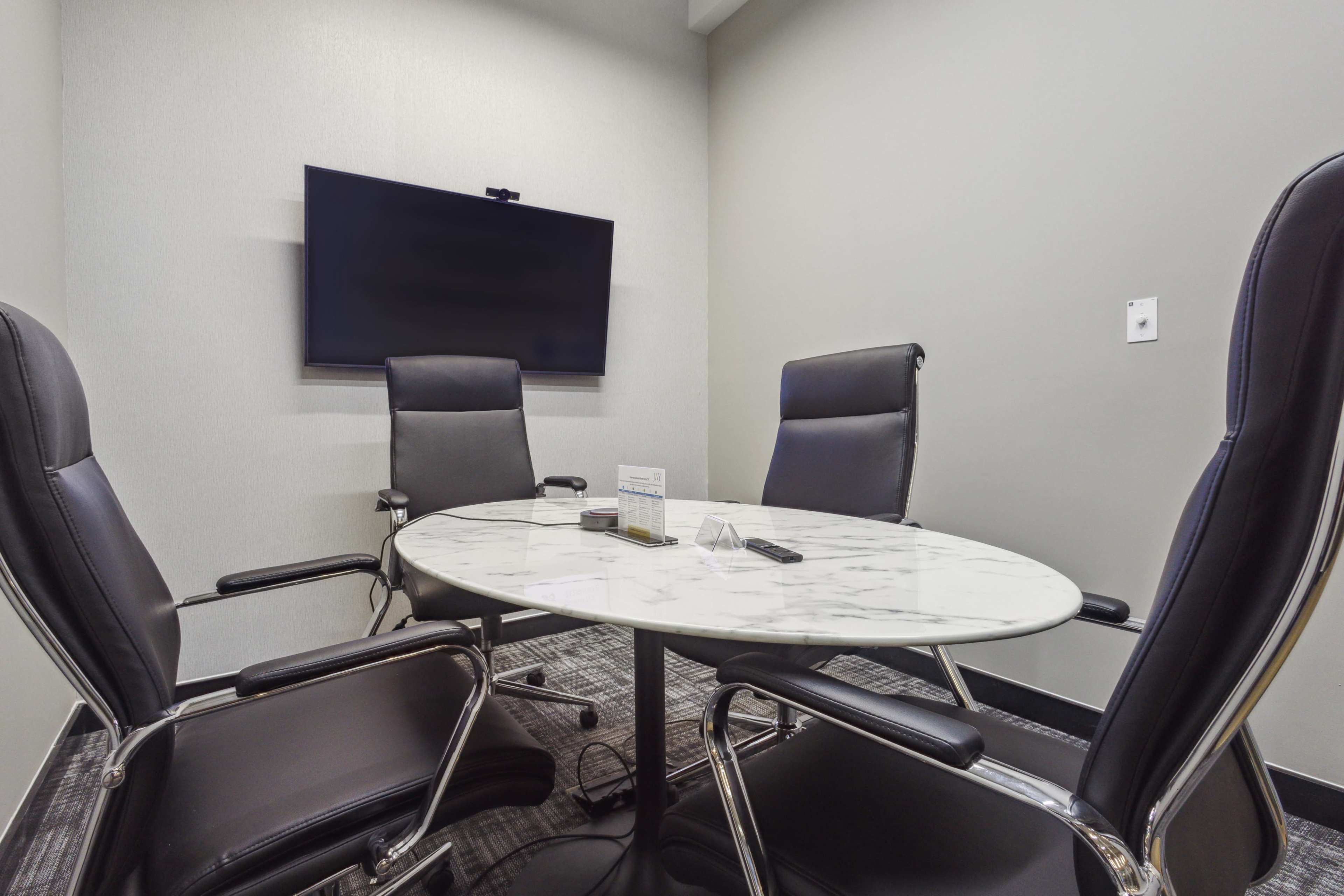 A small meeting room features a marble table surrounded by four black leather chairs and a wall-mounted television.