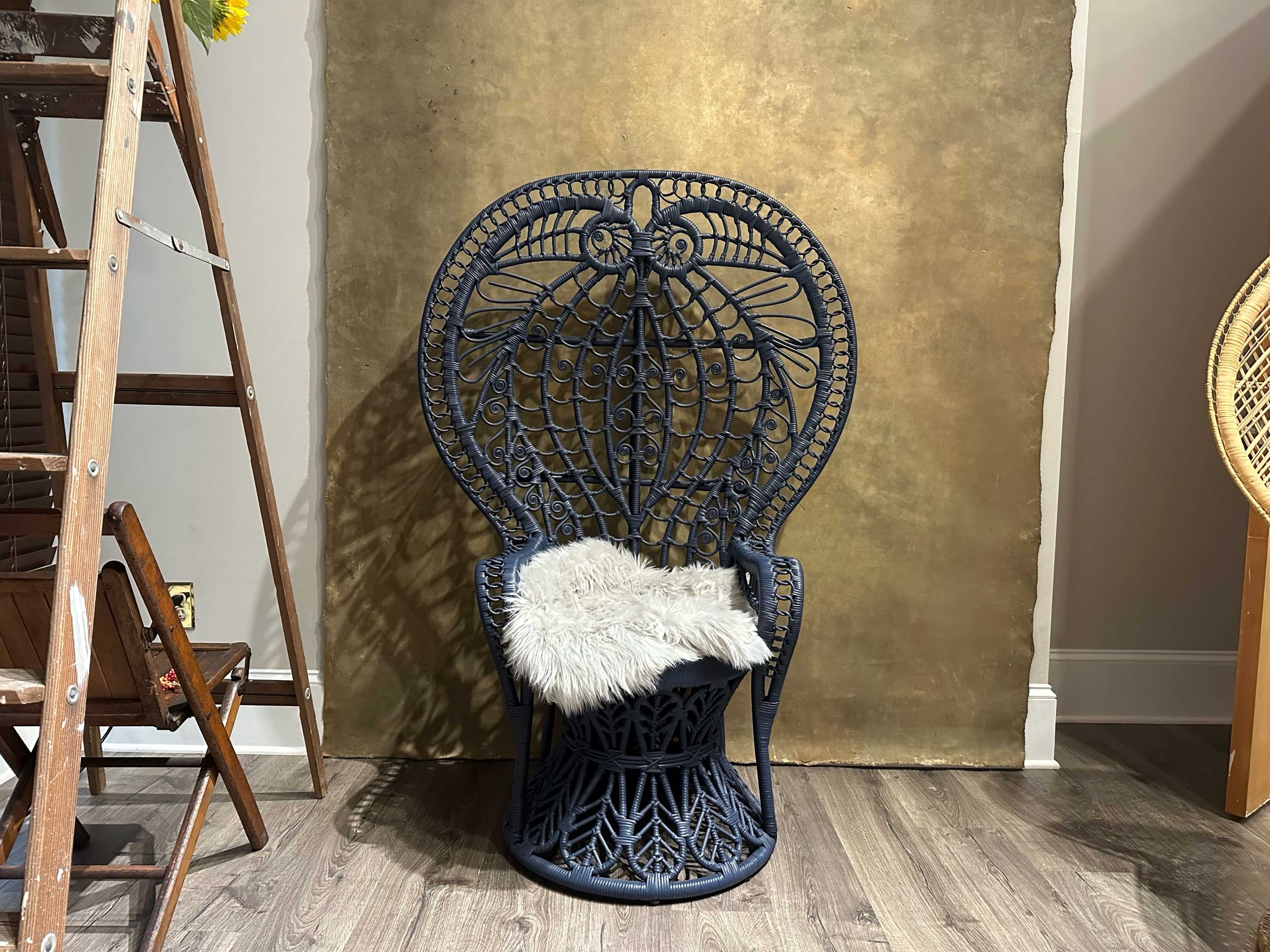 A decorative blue wicker chair with a plush white throw is positioned against a gold backdrop in a room with wooden flooring and a ladder nearby.