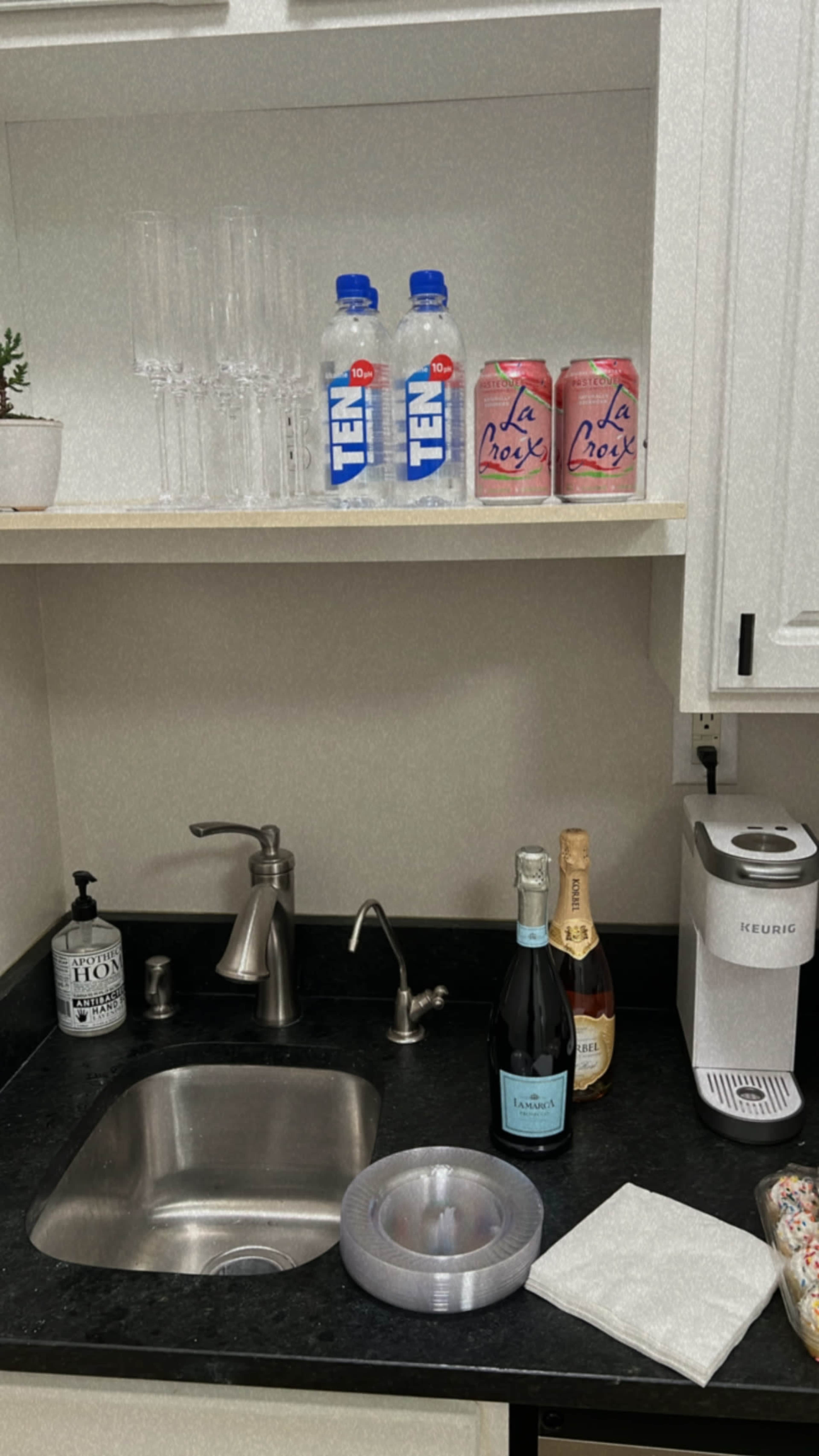 A kitchen counter with a sink, various beverages including water bottles and cans of La Croix, a bottle of champagne, and a coffee machine, along with glasses and a roll of tape.