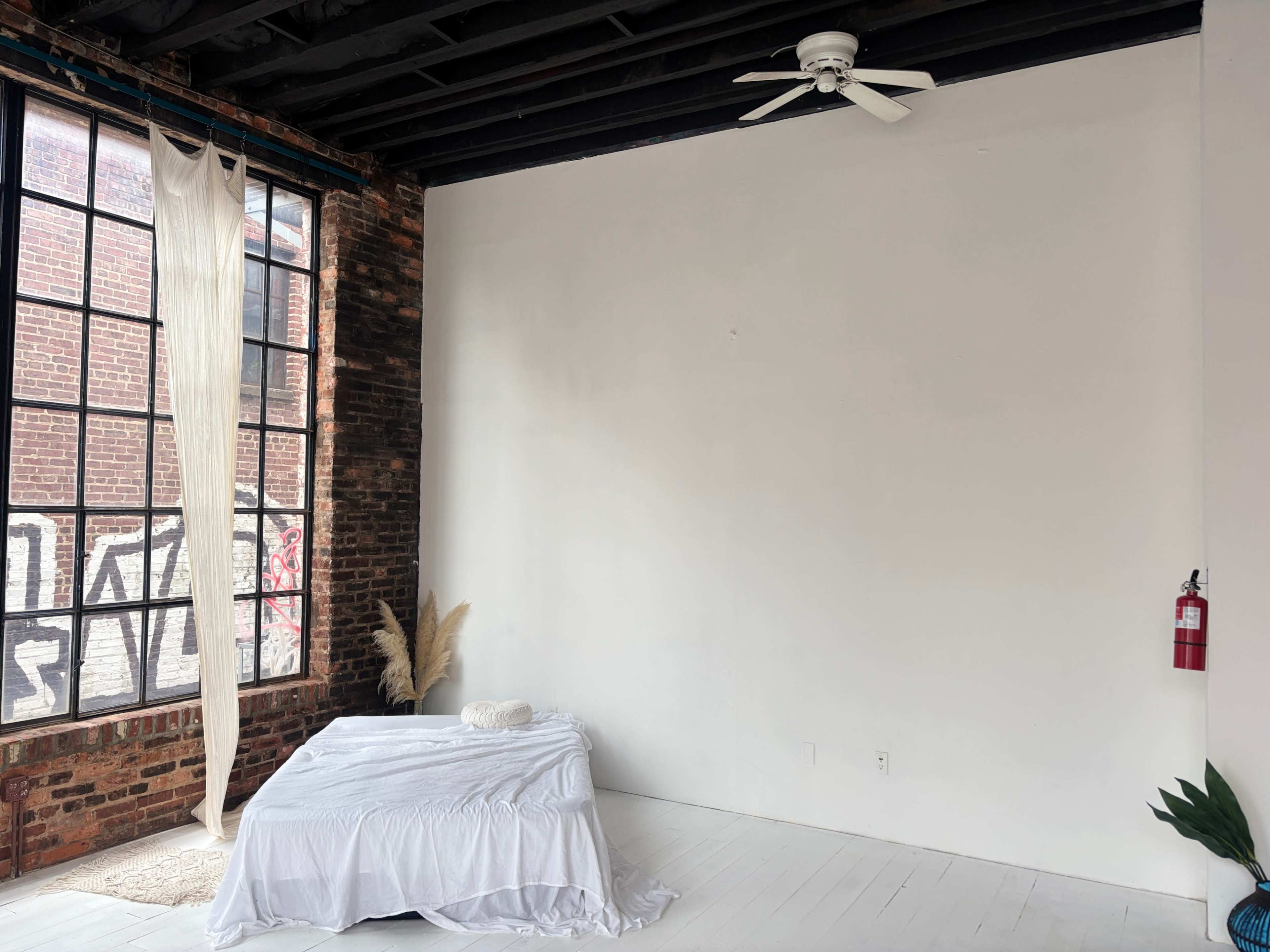 The image shows a minimalist interior space with exposed brick walls, a white bed with a simple covering, and a large window letting in natural light.