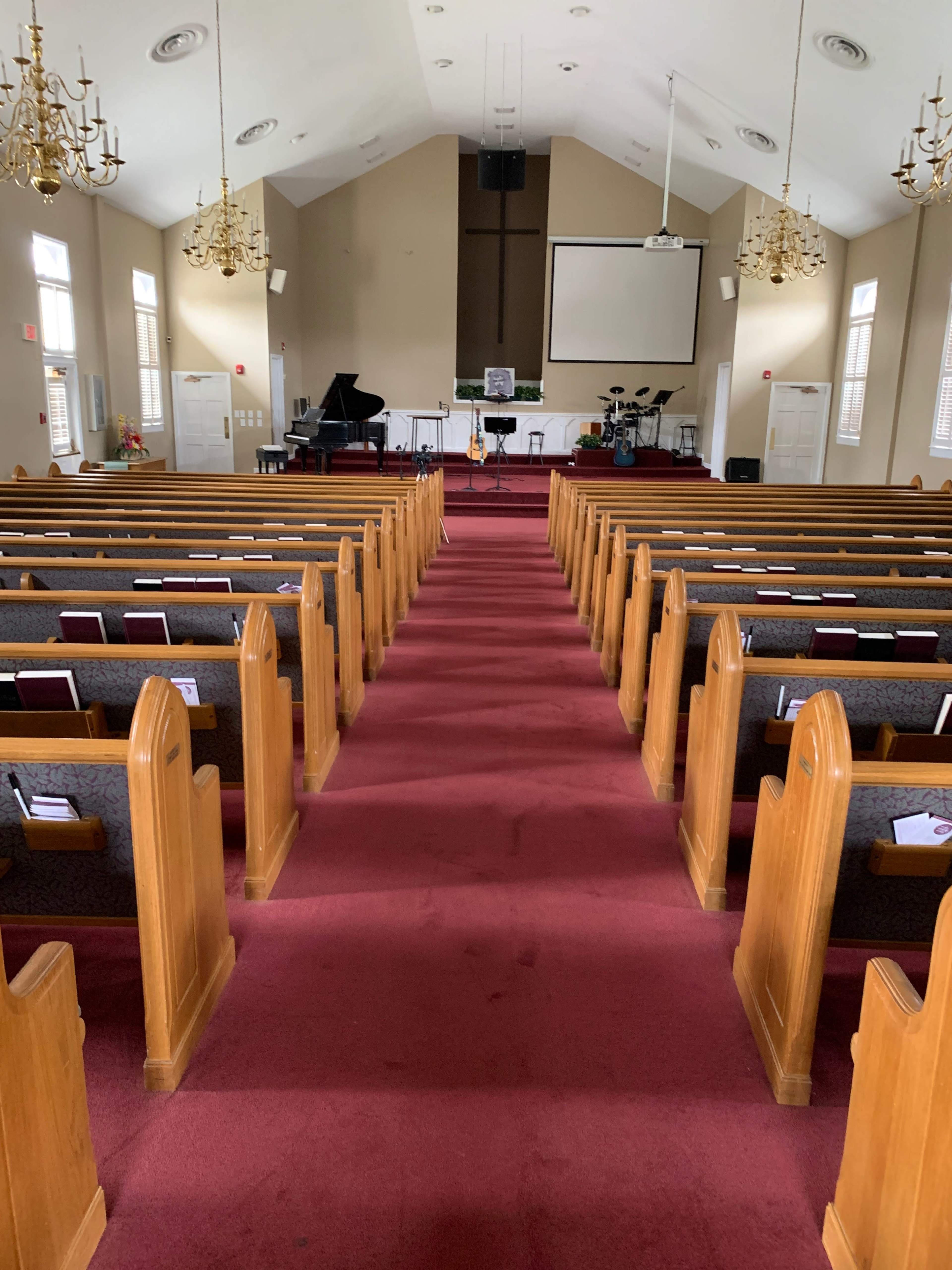 The interior of a church with wooden pews arranged in rows along a red carpet aisle leading to a stage with musical instruments.