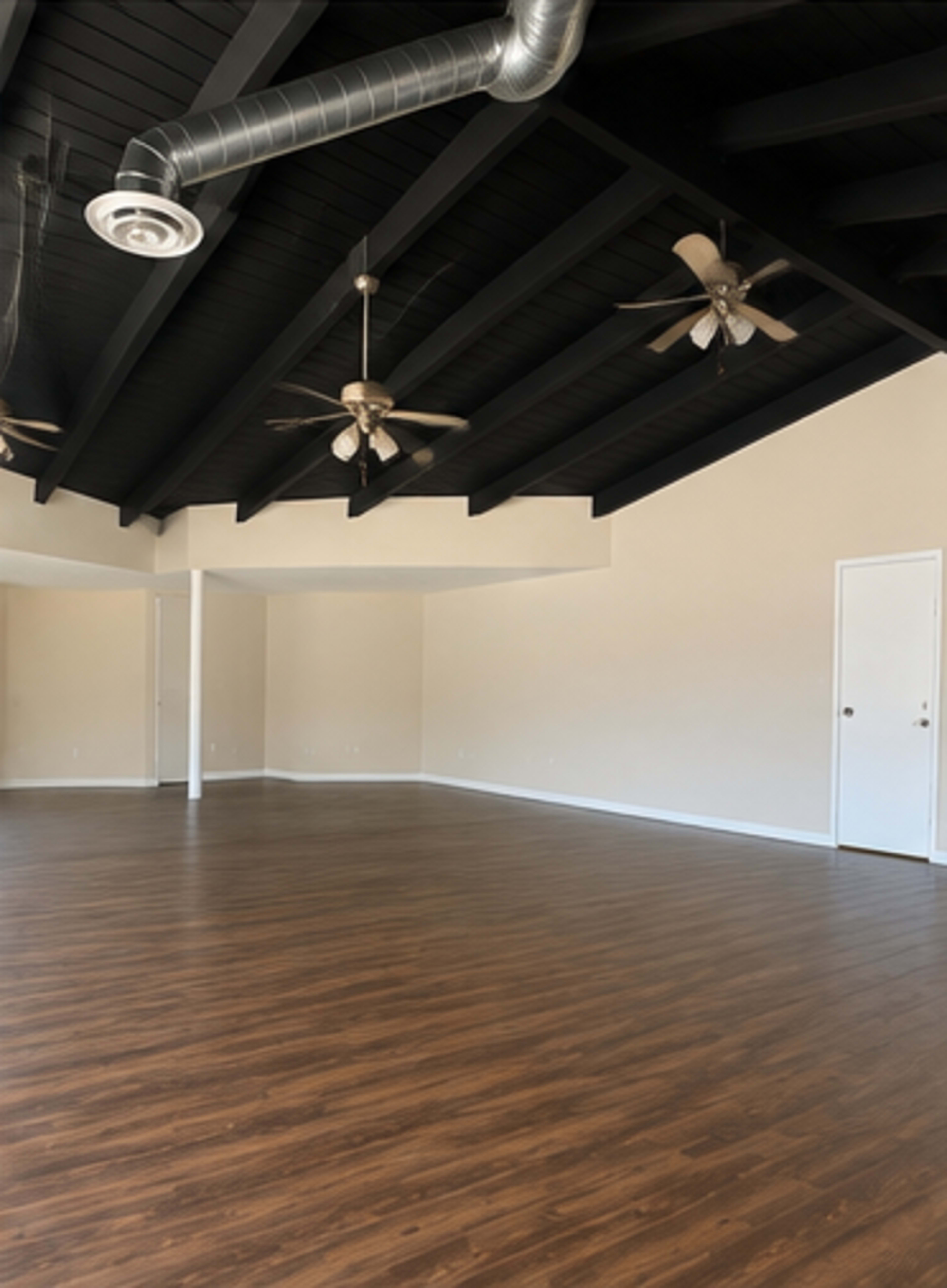 The image shows a spacious room with wooden flooring, high black beam ceilings, and two ceiling fans, along with a door on one wall.