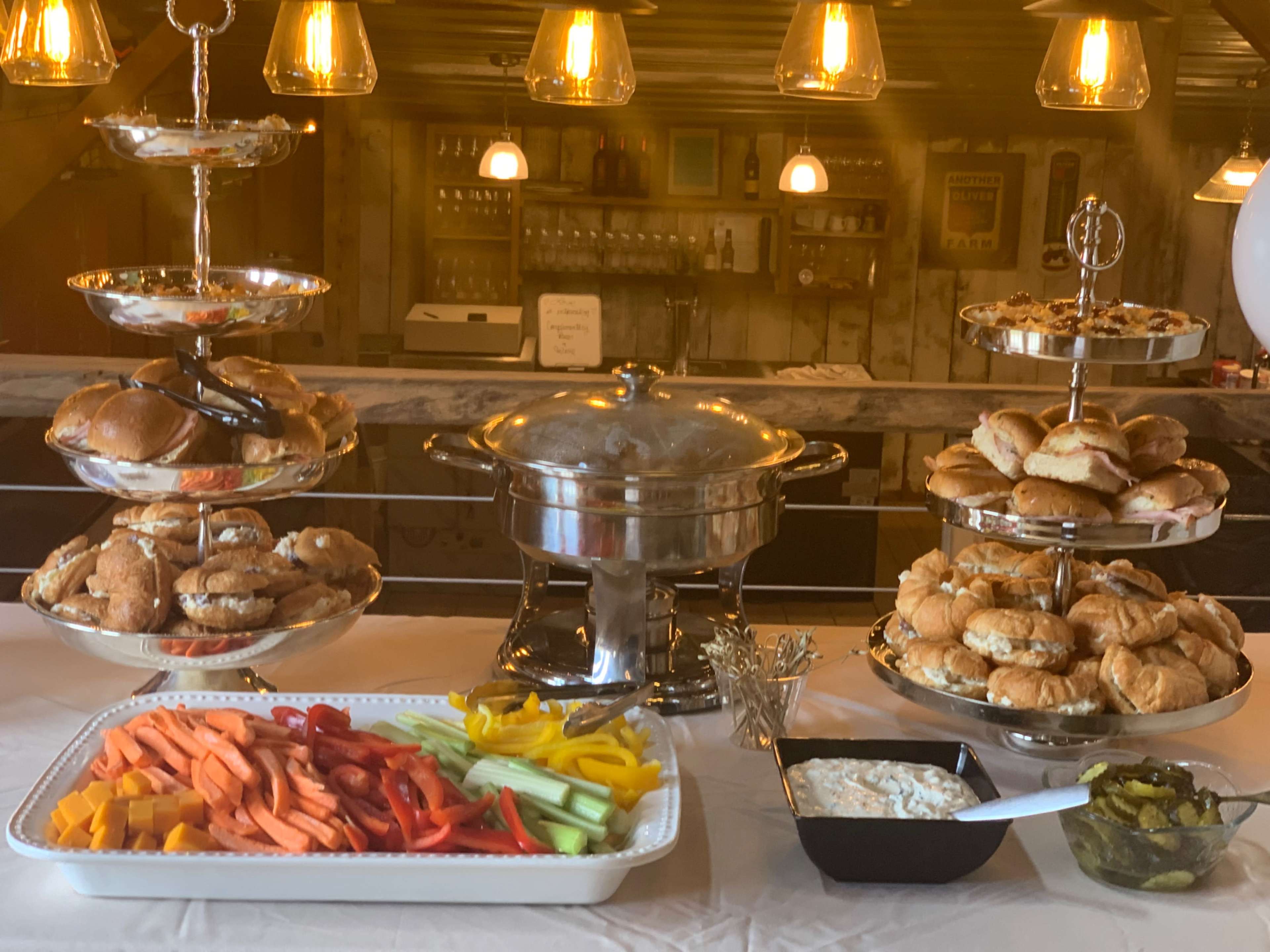 A buffet setup features two tiers of assorted sandwiches and pastries, a chafing dish with hot food, and a platter of fresh vegetables and dip.