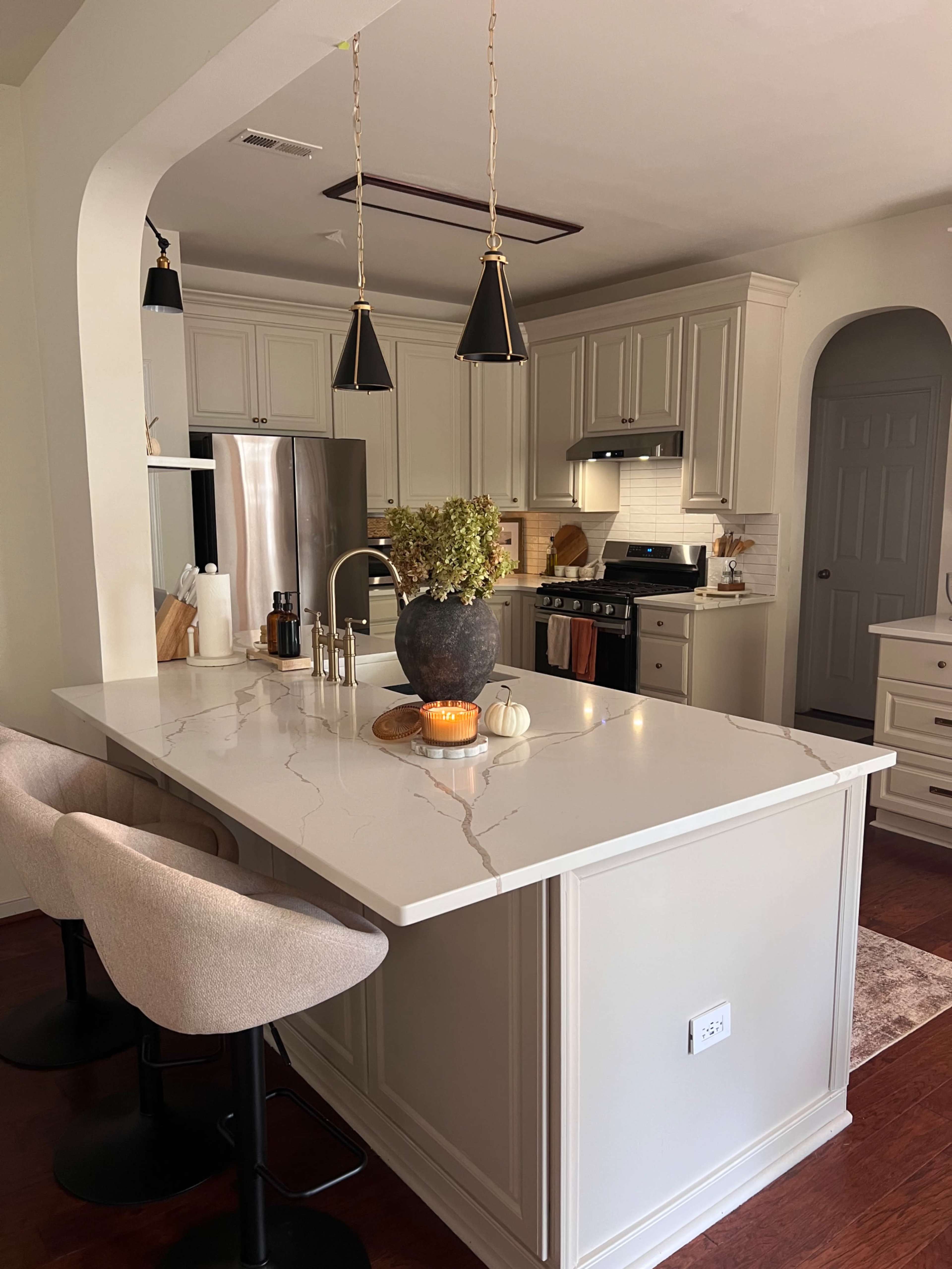 The image shows a modern kitchen featuring a large island with a marble countertop, two bar stools, stainless steel appliances, and warm lighting from pendant fixtures.