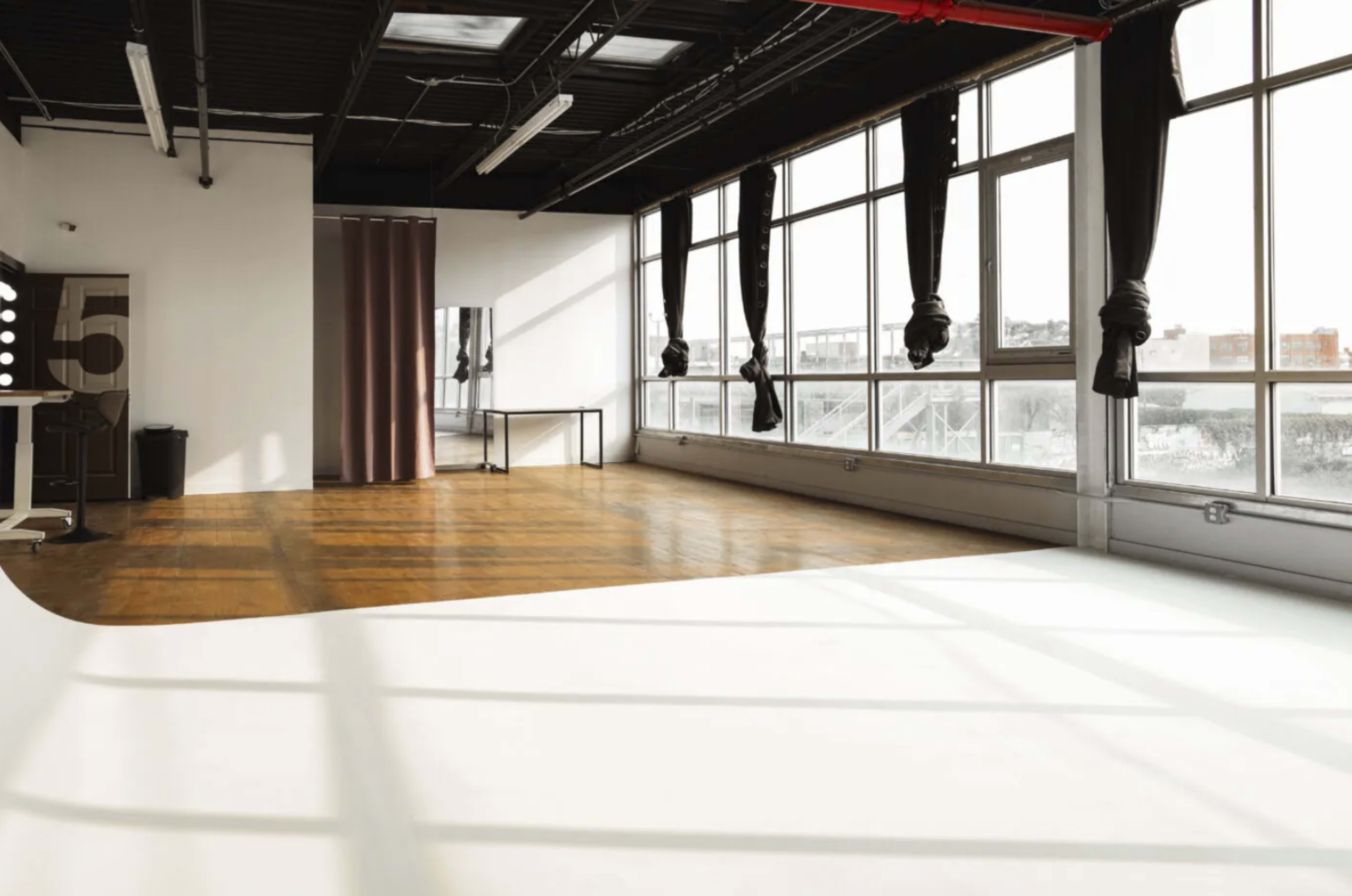 A spacious, well-lit studio with large windows, hardwood floors, and a white backdrop.