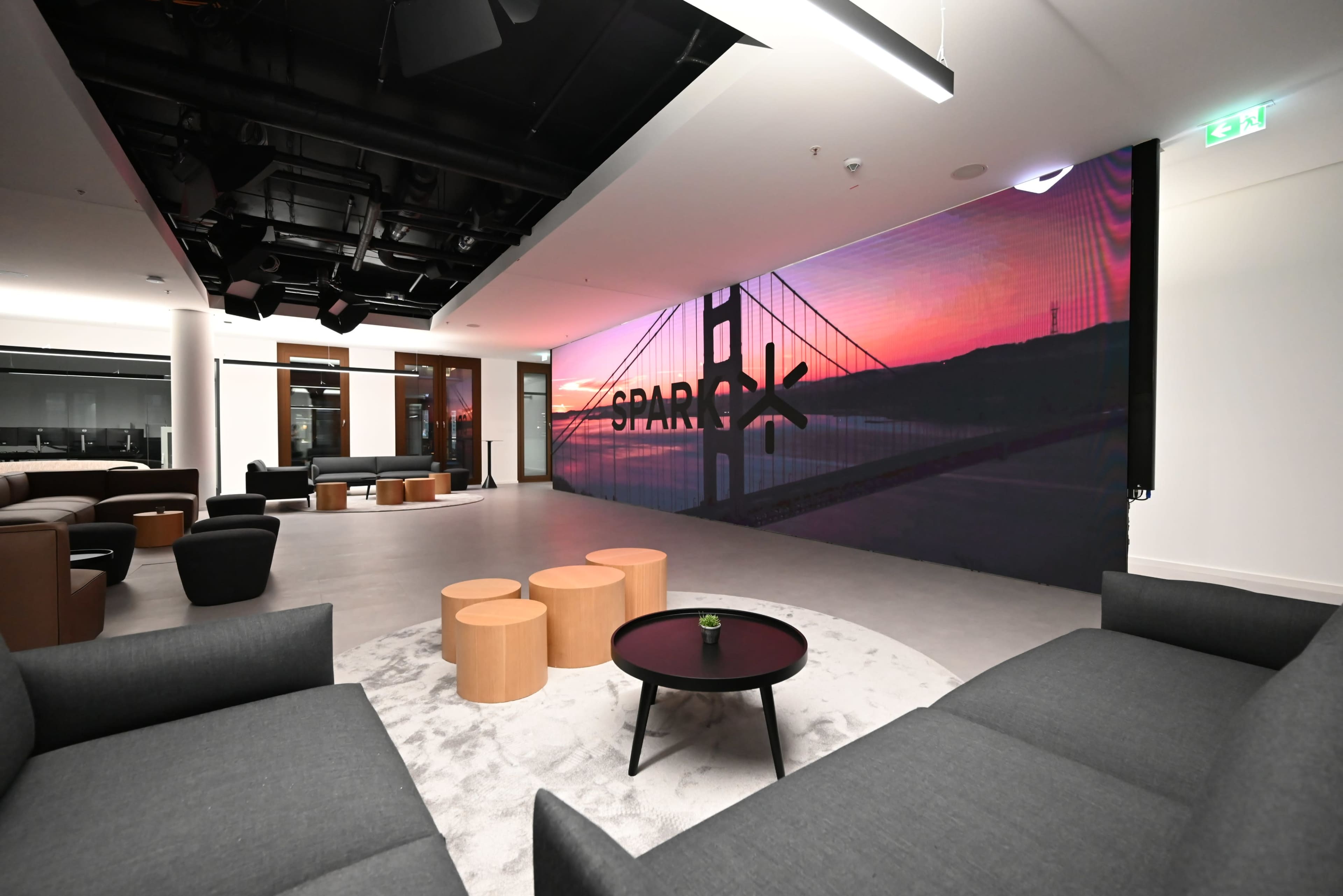 A modern lounge area with gray sofas, wooden coffee tables, and a large digital display featuring a sunset over a bridge.
