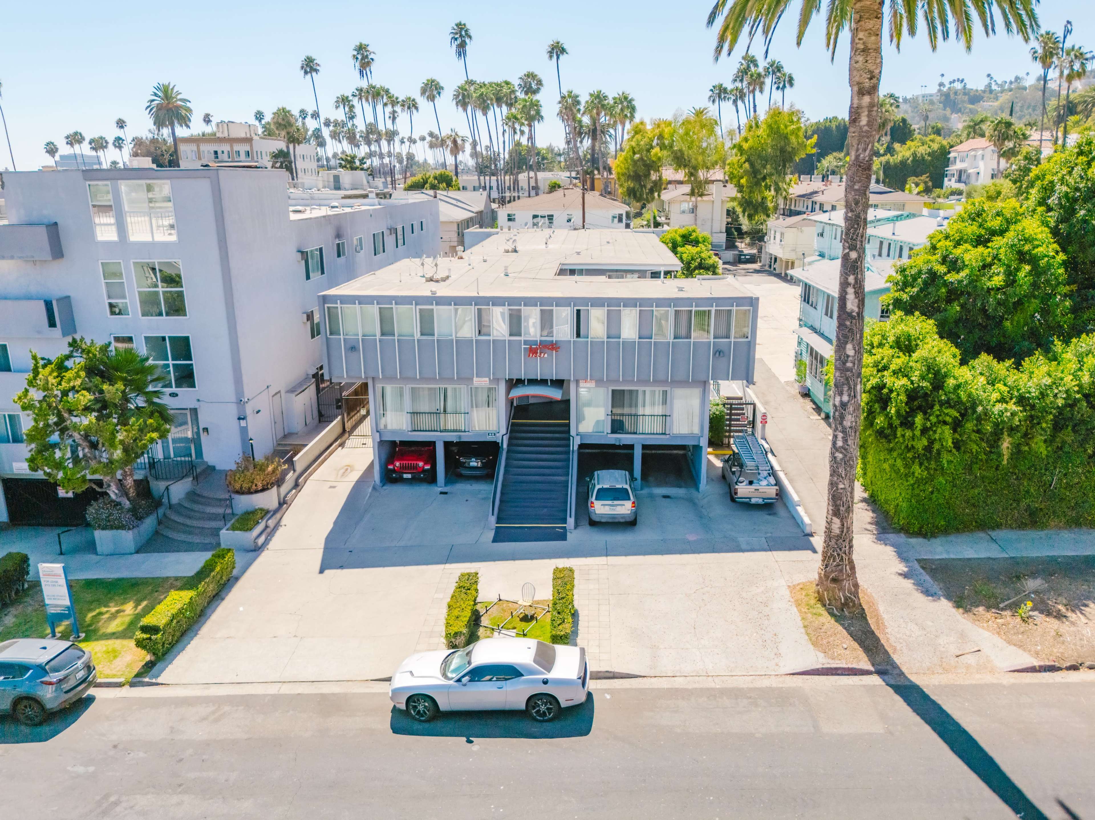 The image shows a two-story apartment building with palm trees in the background, parked cars in front, and a well-maintained lawn area.