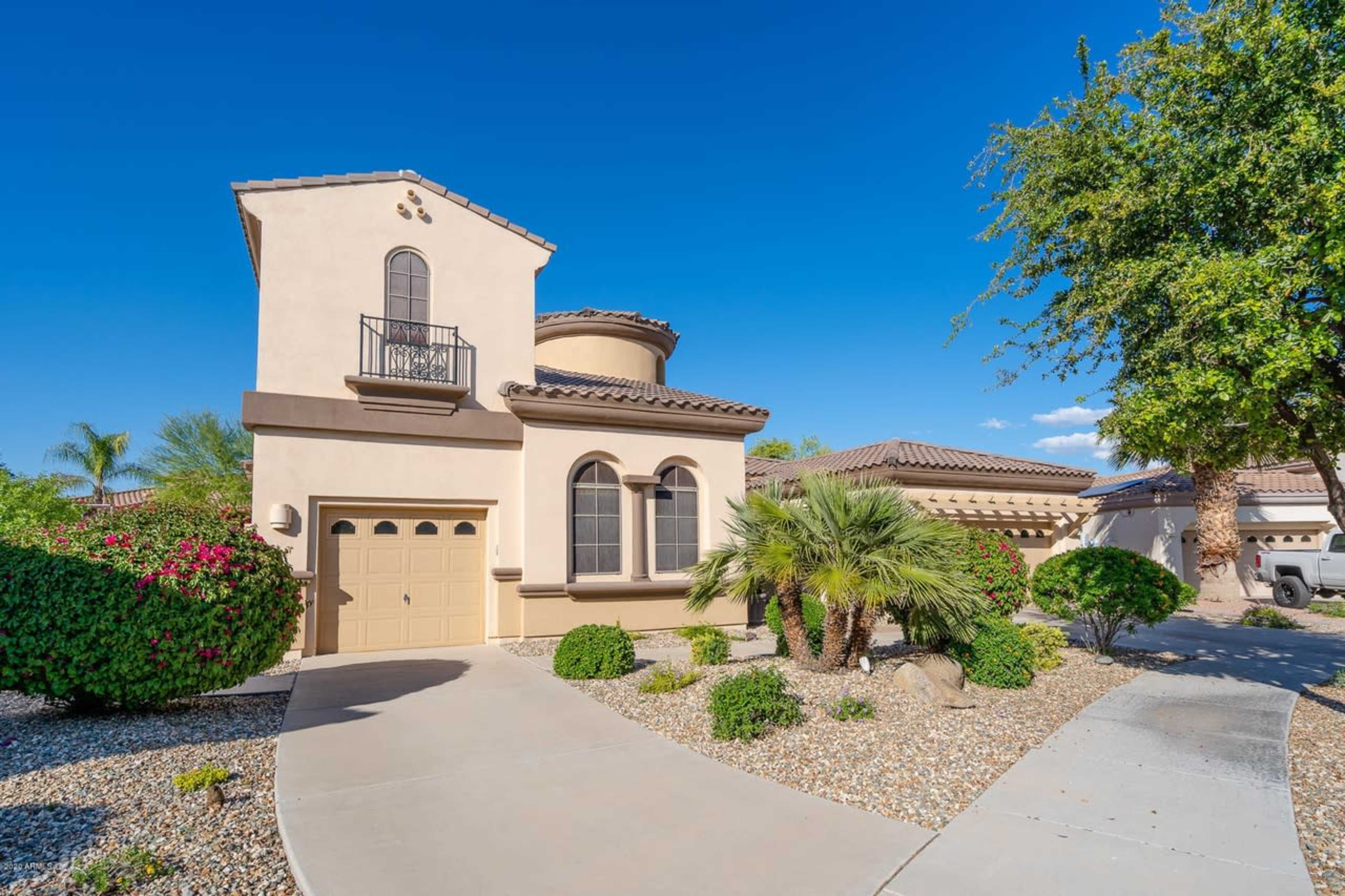 A two-story beige house with a round turret, garage, and landscaped front yard featuring palm trees and decorative rocks.