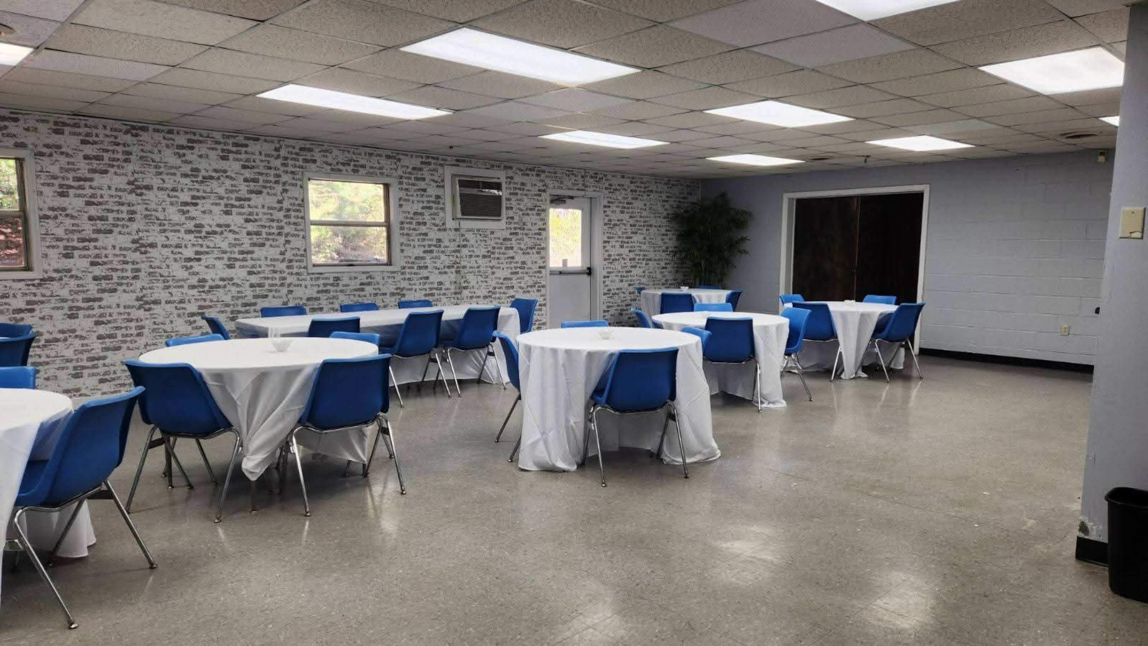 The image shows a large, empty room with several round tables covered in white tablecloths and blue chairs arranged around them, along with a patterned brick wall and large windows.