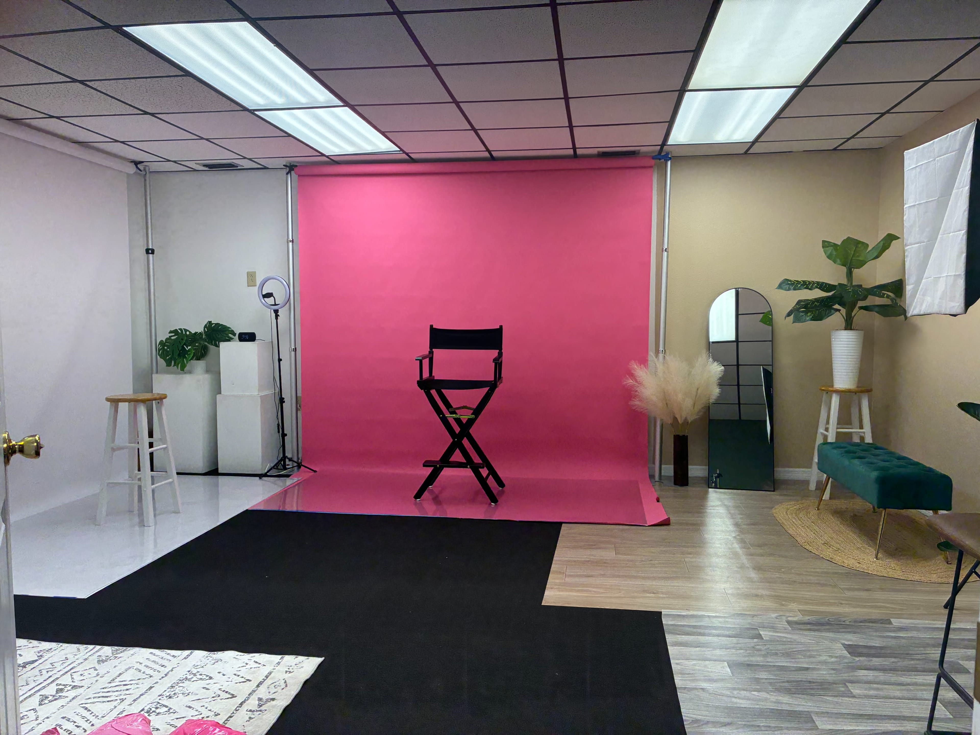 The image shows a studio space with a pink backdrop, a black director's chair in front, and various decorative elements including plants and seating arrangements.