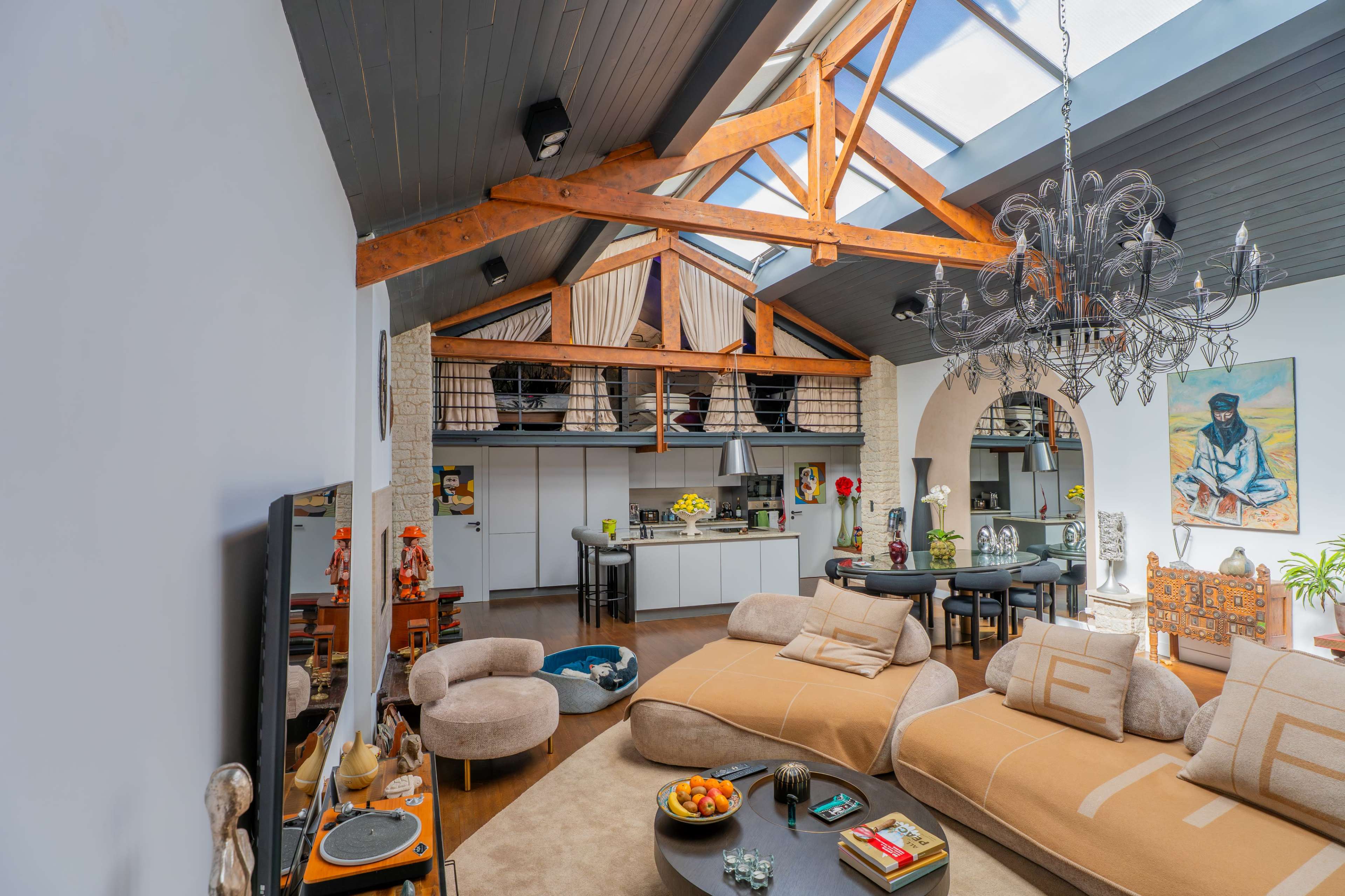 The image shows a spacious, modern living room with exposed wooden beams, a chandelier, and a cozy seating area, featuring a kitchen and dining space in the background.