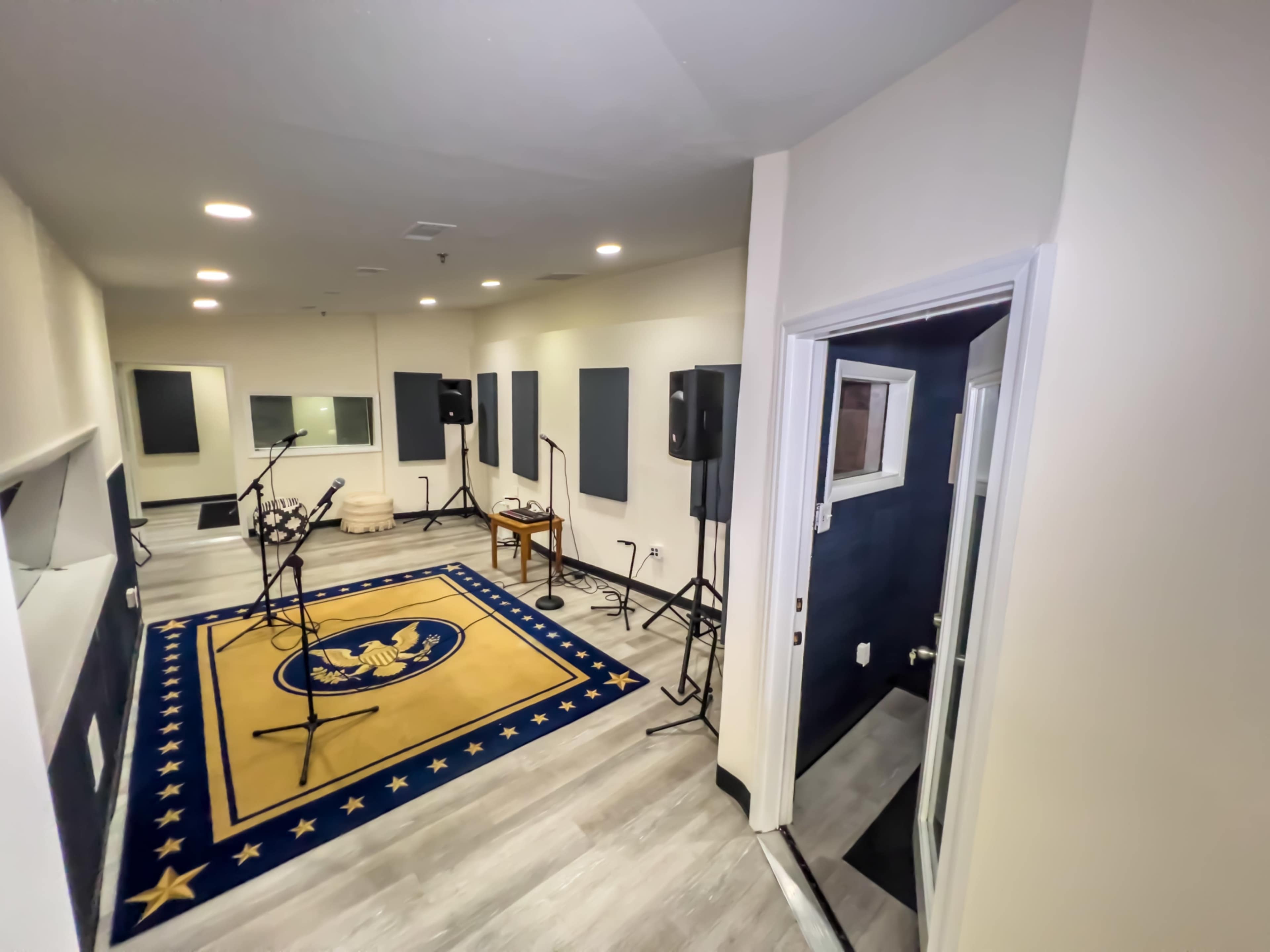 A recording studio featuring a large area with microphones on stands, gray soundproofing panels on the walls, and a rug with an emblem in the center.