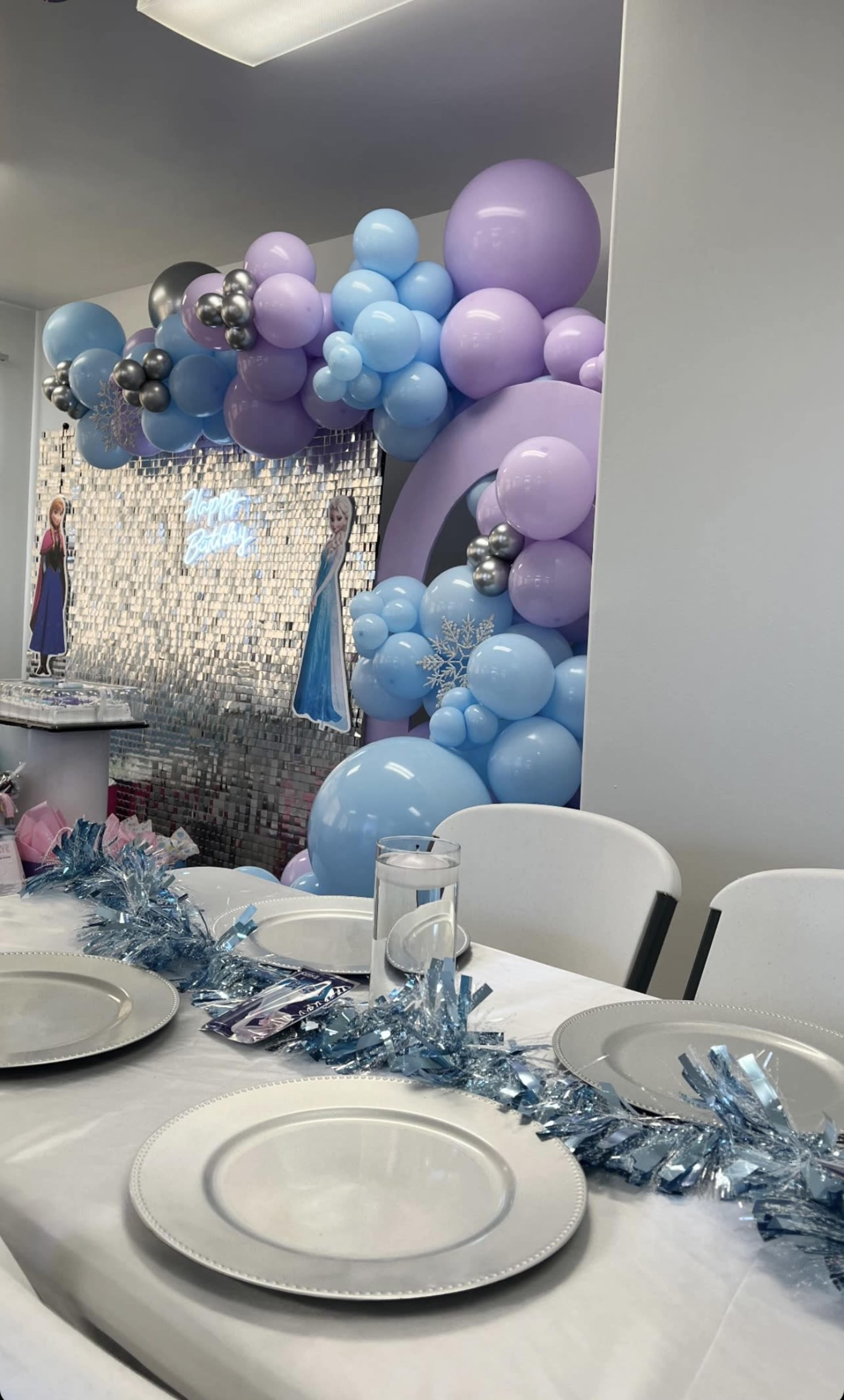 A birthday party setting features a table with plates and a decorative light blue and purple balloon backdrop, alongside figures resembling ice-themed characters.