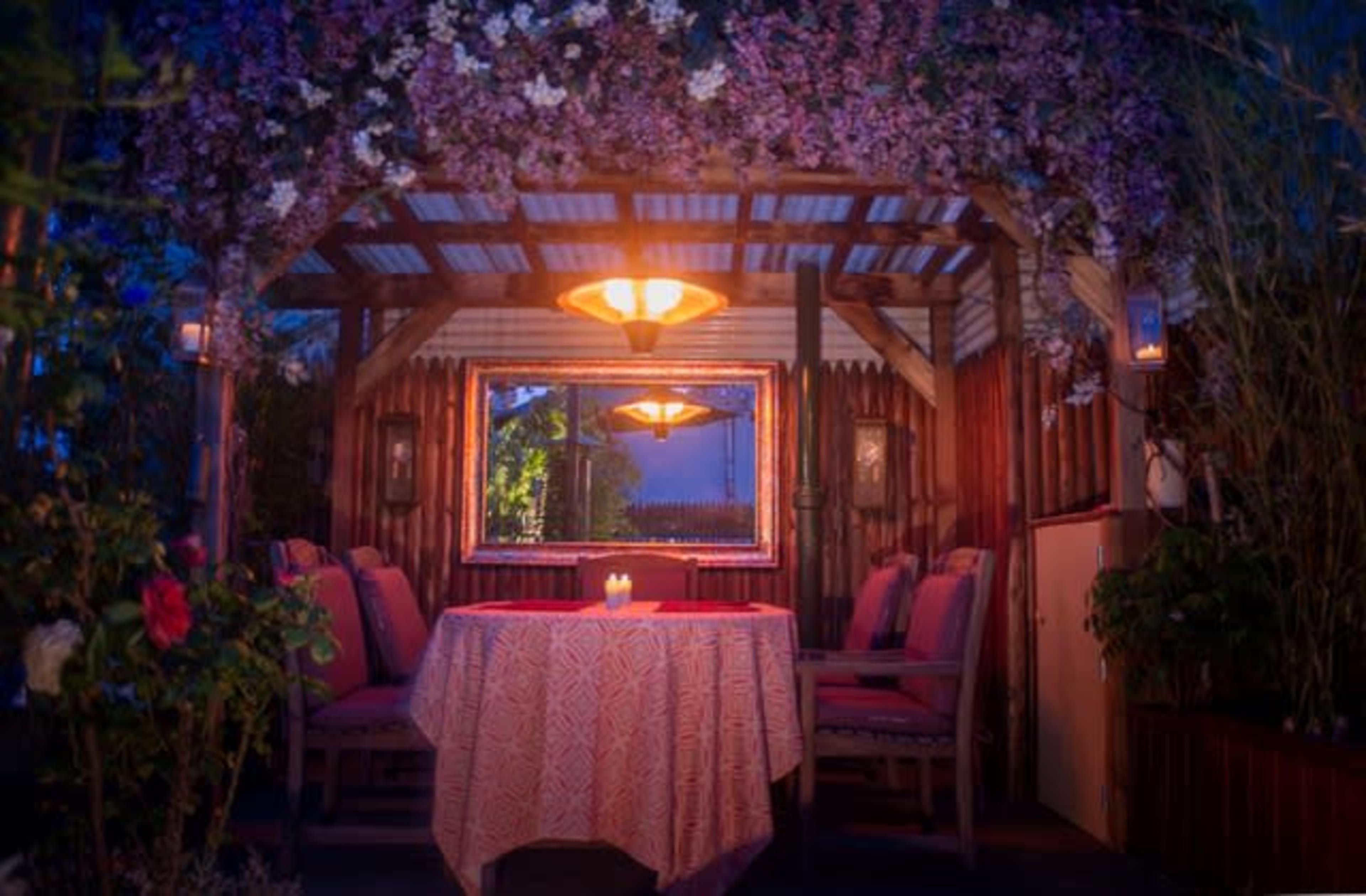 A cozy outdoor dining space features a table covered with a patterned cloth, surrounded by chairs, and is adorned with hanging flowers and a mirror under soft lighting.