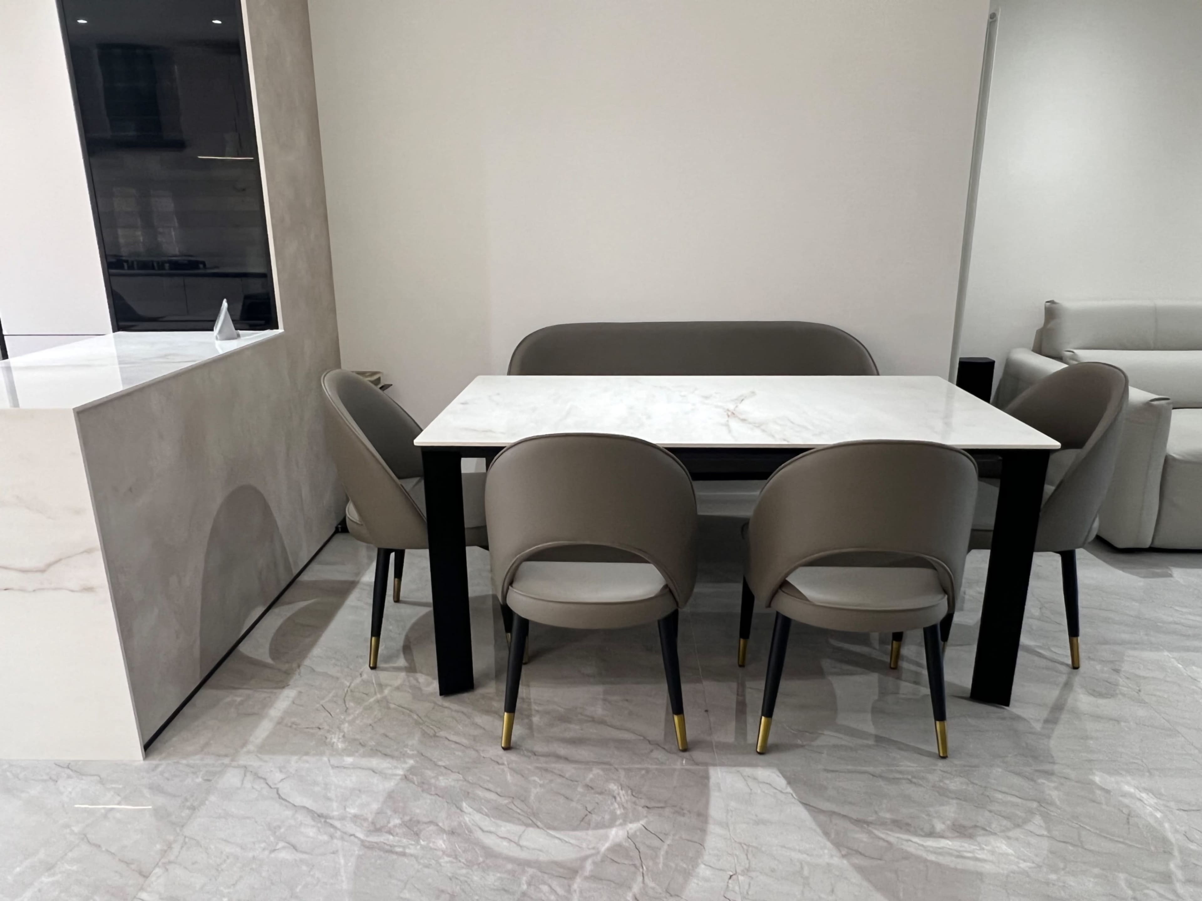 A marble-top dining table with four beige chairs is positioned in a modern interior space with light-colored flooring.