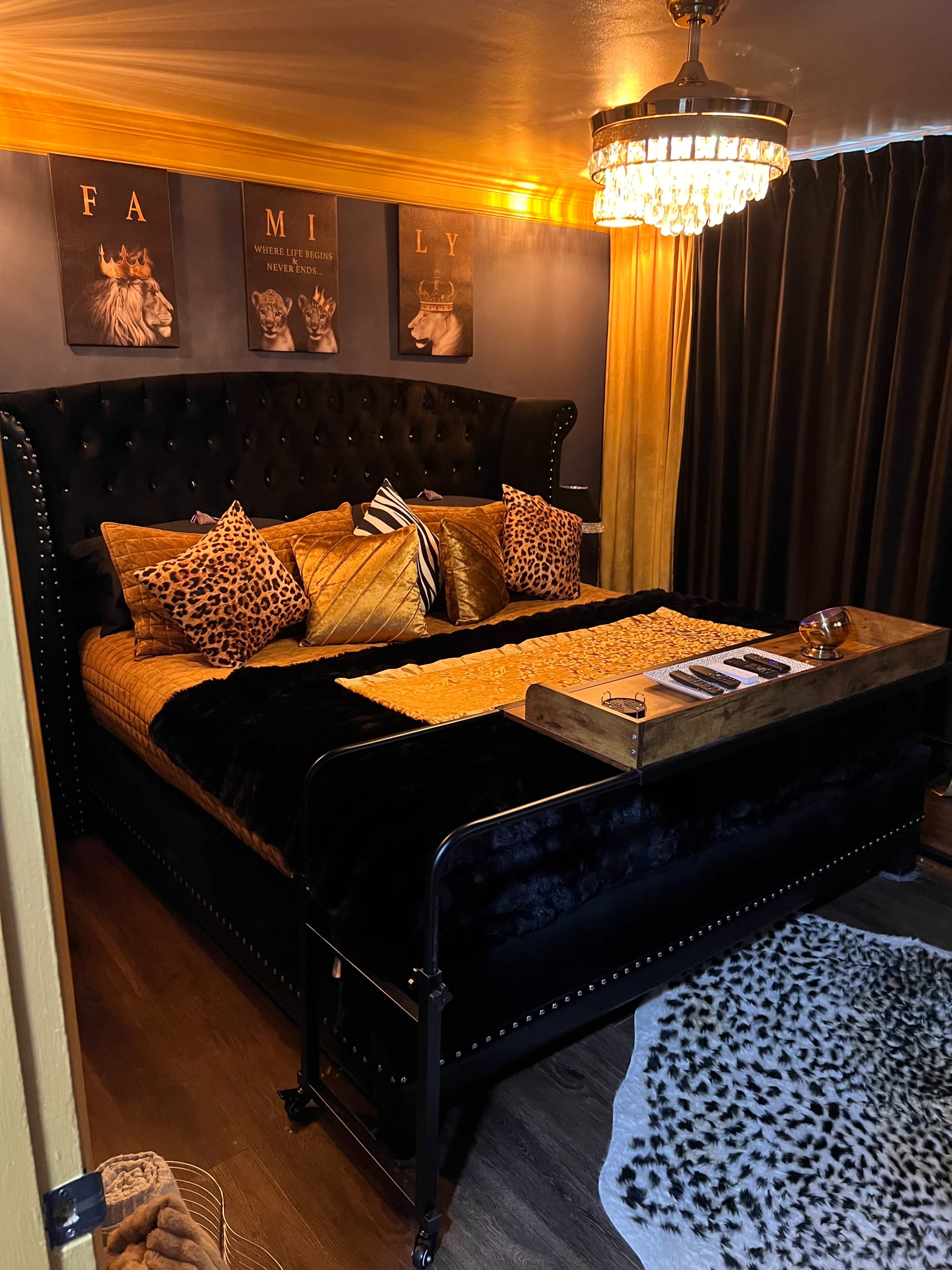 The image shows a luxurious and modern bedroom featuring a black tufted bed covered with patterned pillows and a vibrant yellow throw, surrounded by dark curtains and a chandelier overhead.