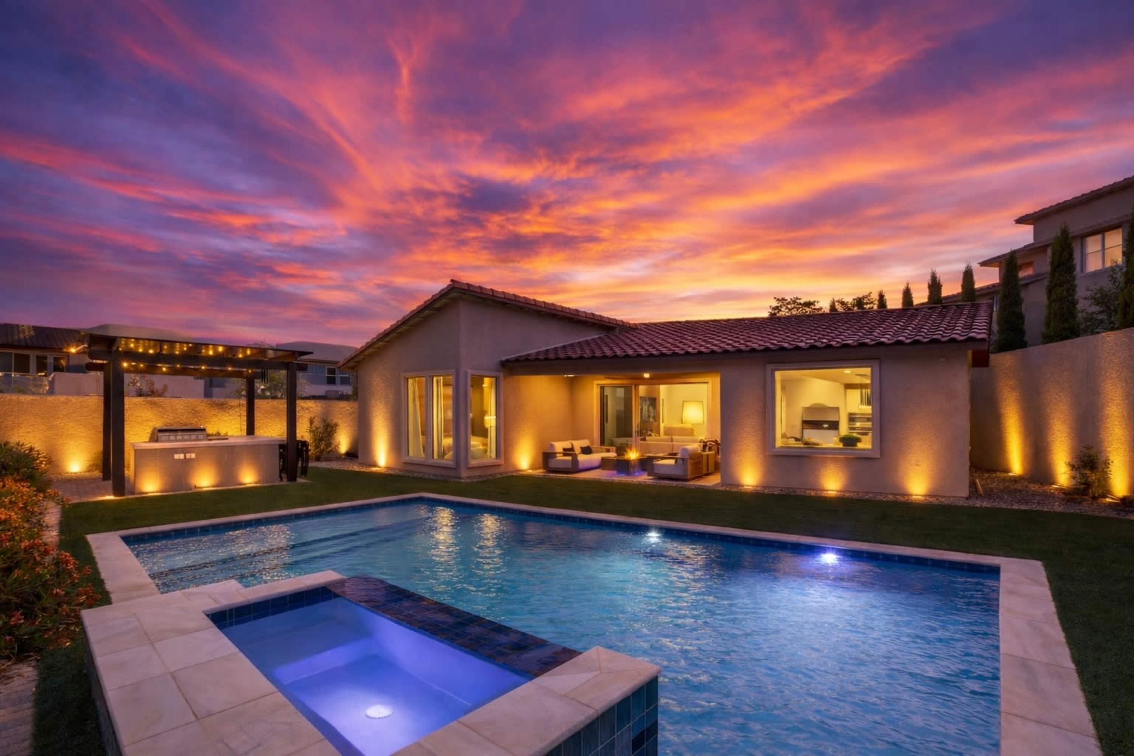A modern house with a swimming pool is illuminated against a vibrant sunset sky, featuring a landscaped yard and patio area.