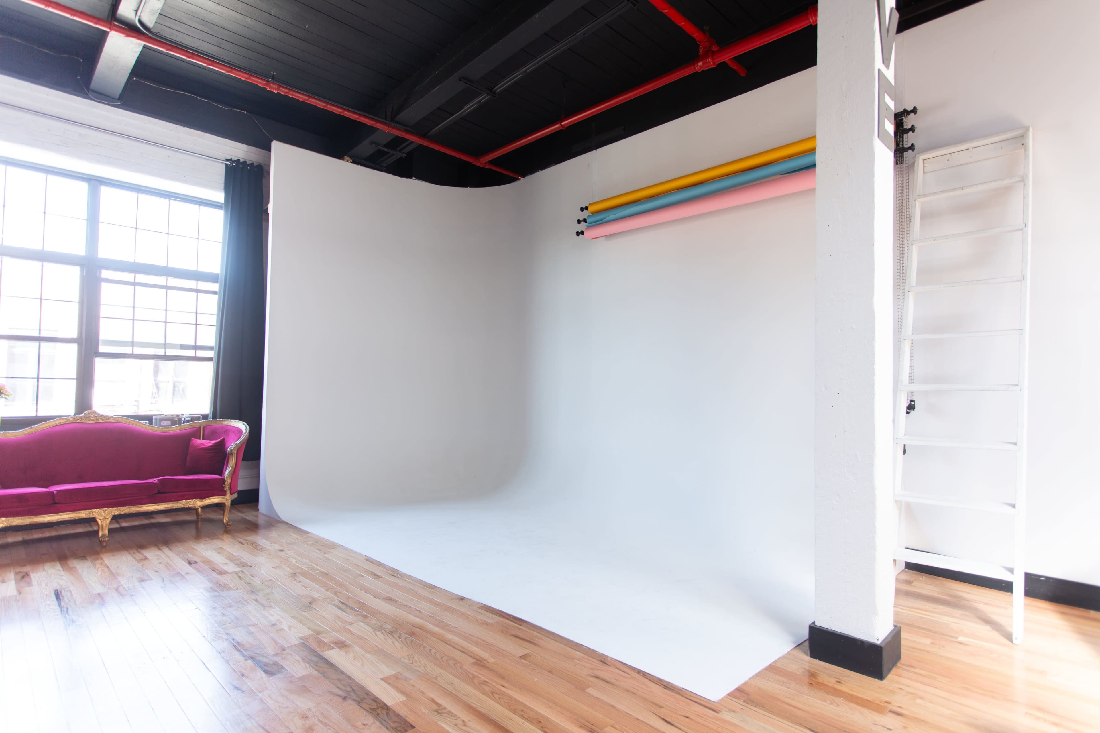 The image shows a photography studio with a curved white backdrop, colorful backdrop paper rolls, and a pink velvet sofa against a large window.