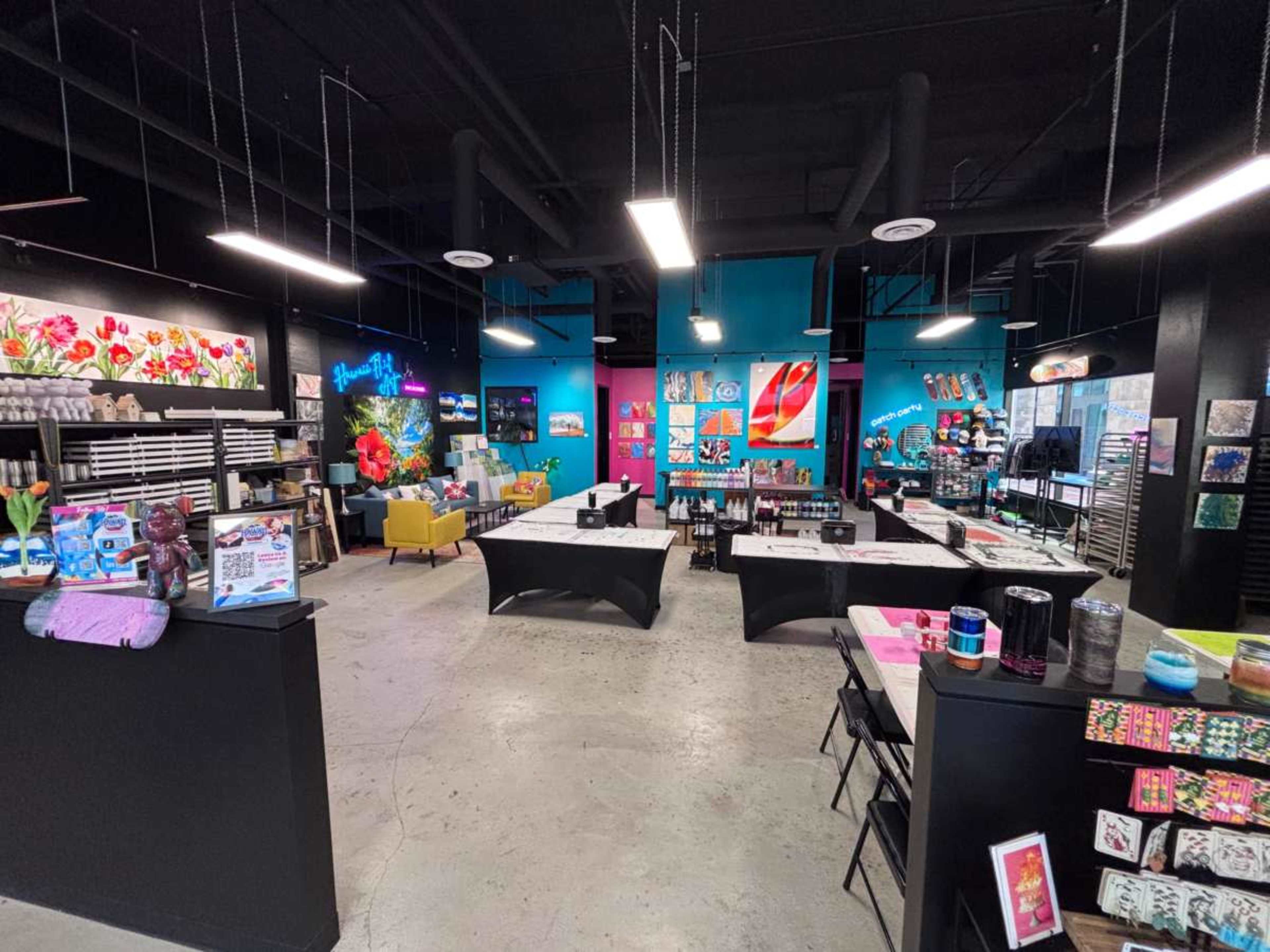 The image shows a vibrant art studio or creative space featuring colorful artworks on the walls, tables set up for activities, and various art supplies displayed for sale.