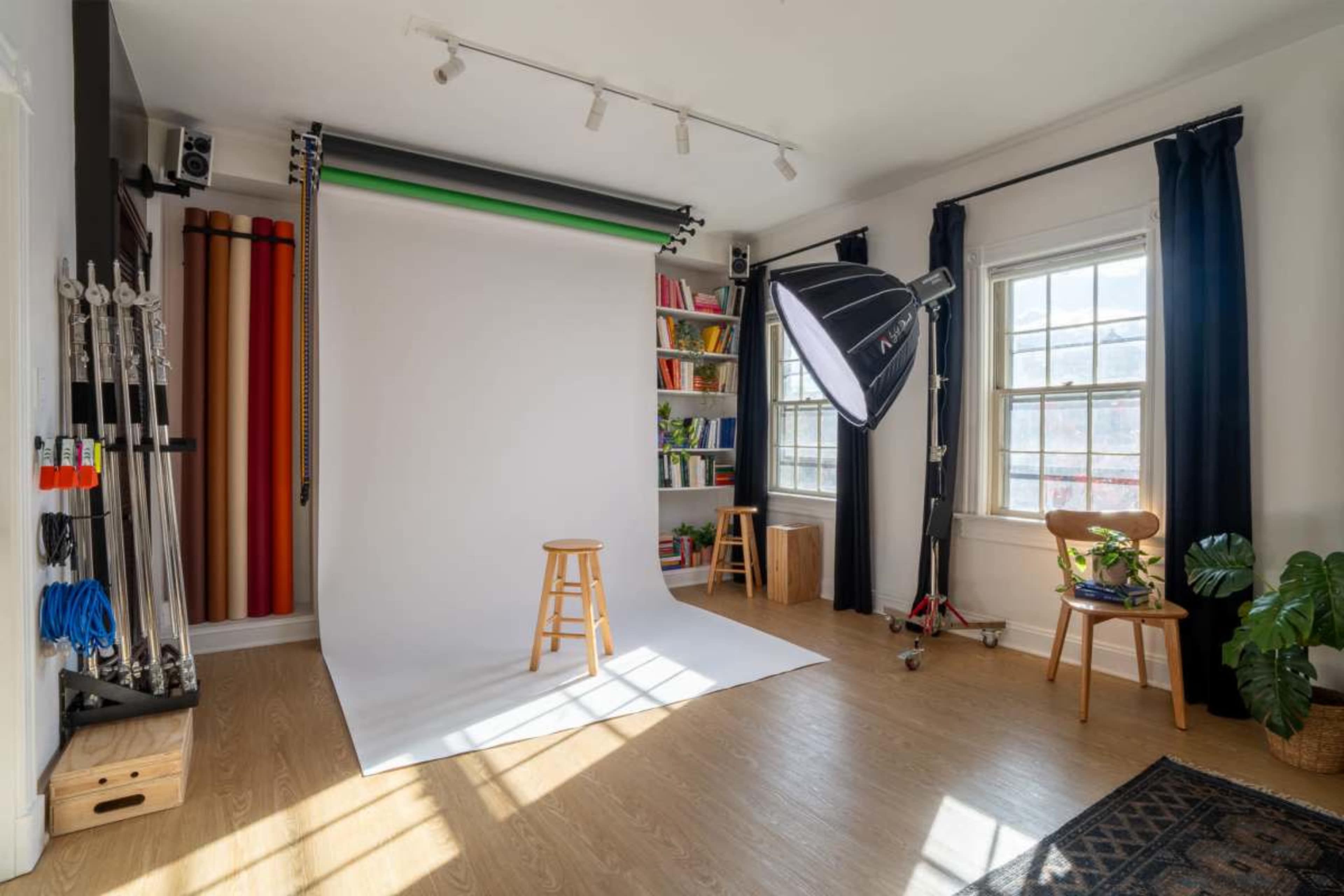 The image shows a well-lit photo studio featuring a white backdrop, a wooden stool, and a large light source, with color backdrops and a bookshelf visible in the background.