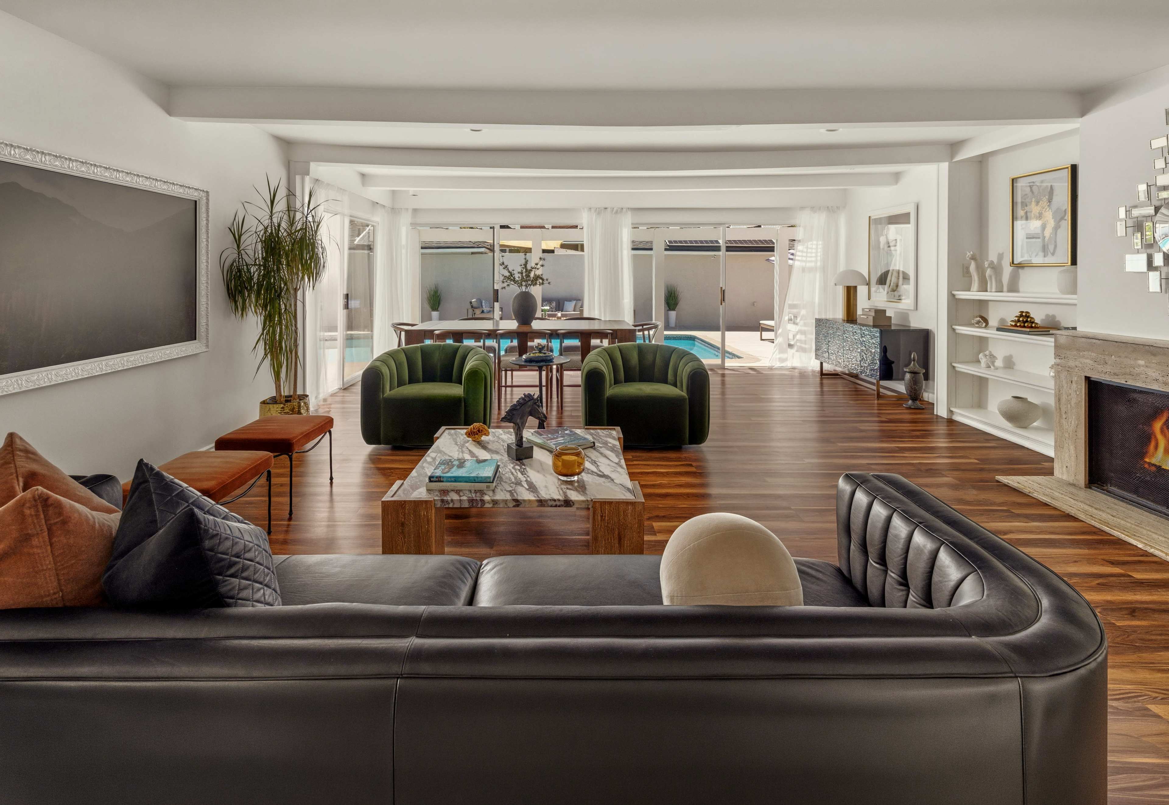 The image shows a modern, open-concept living room with a black sectional couch, green accent chairs, and a view of a pool area through large sliding glass doors.