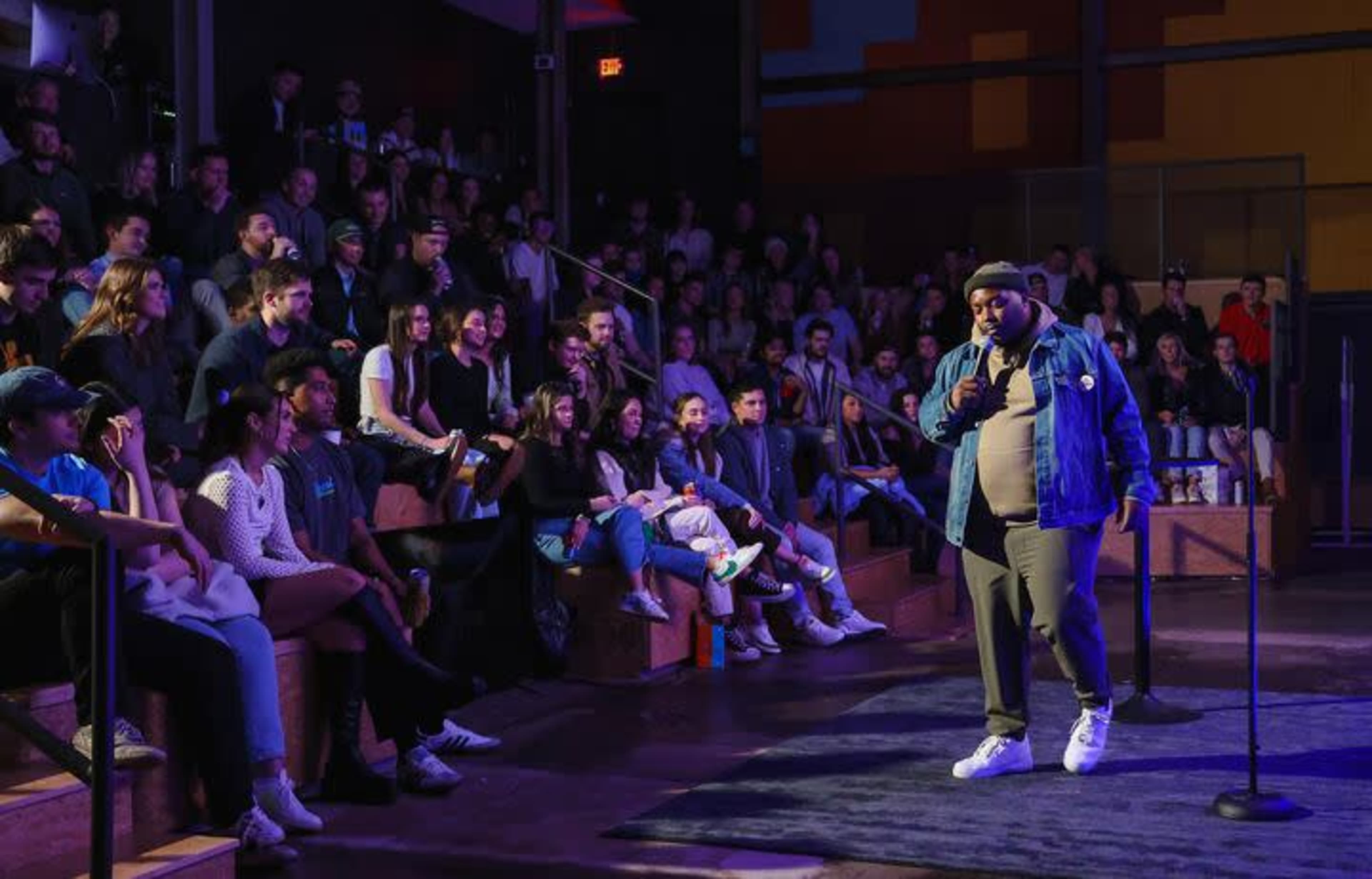 A comedian performs on stage in front of an audience seated in a dimly lit venue.