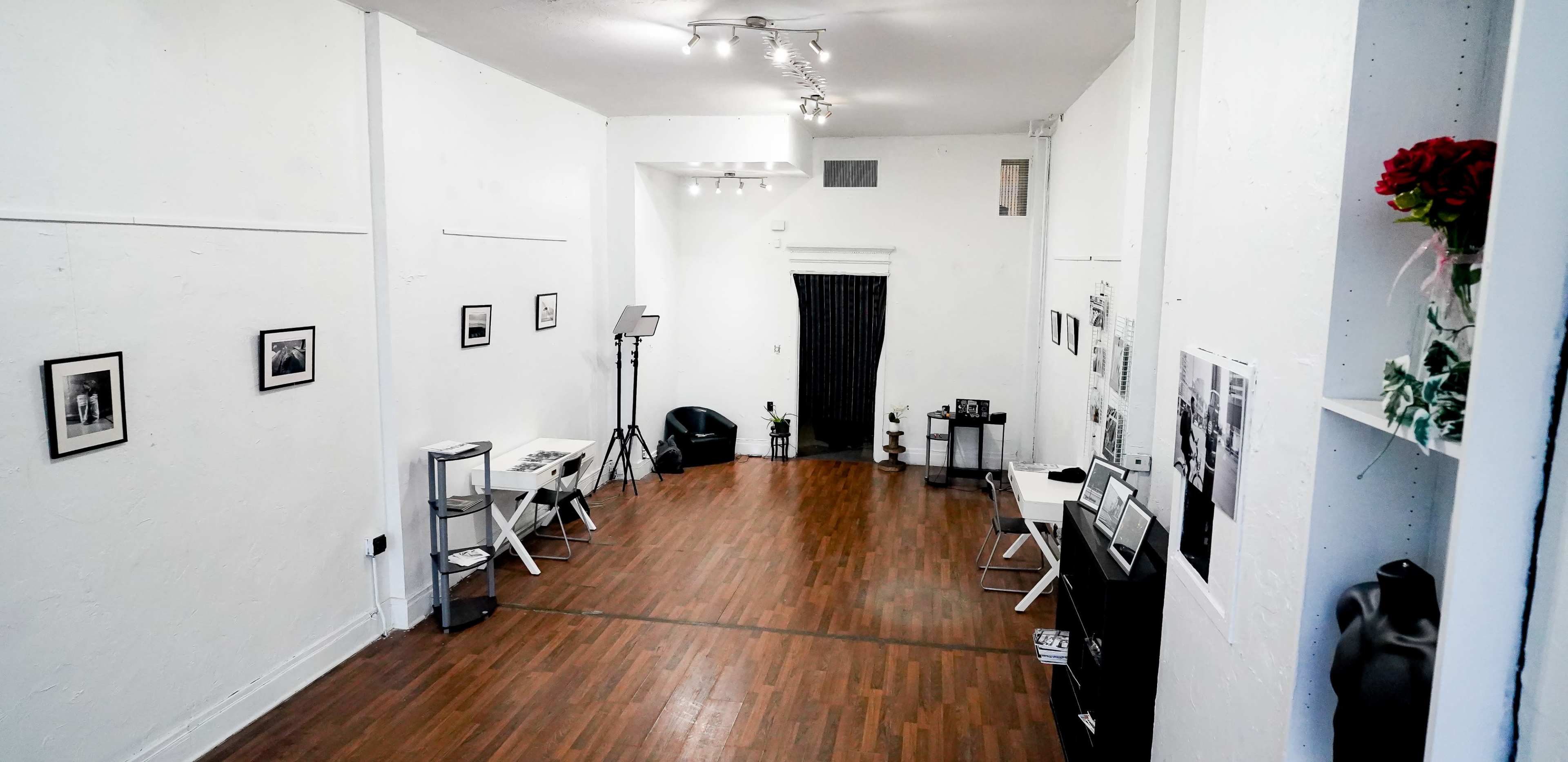 A spacious, minimally furnished room with wooden flooring, white walls, and several black-and-white photographs displayed on the walls.