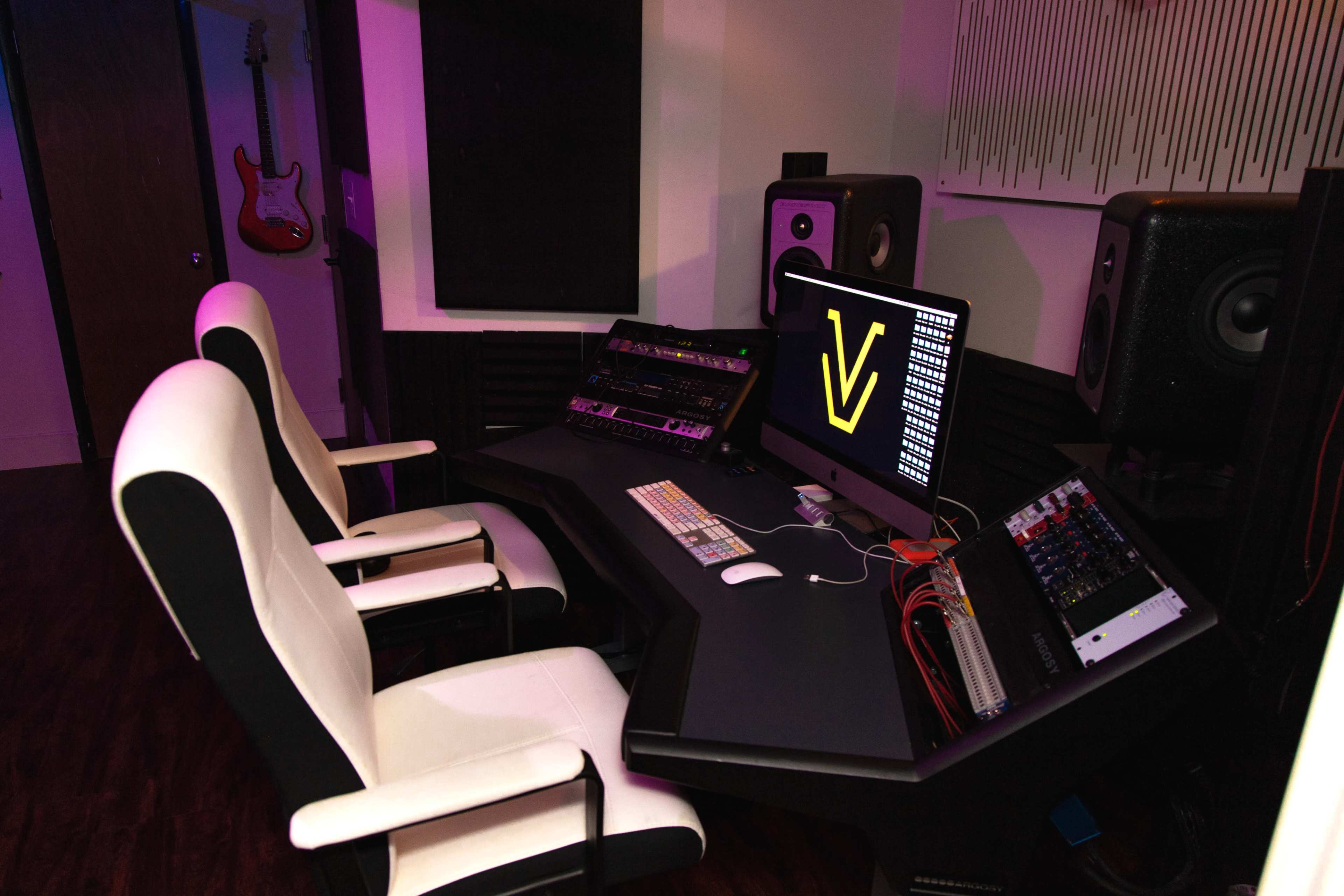 A modern music studio with a sleek black desk, two ergonomic white chairs, computer equipment, and two audio speakers on either side.