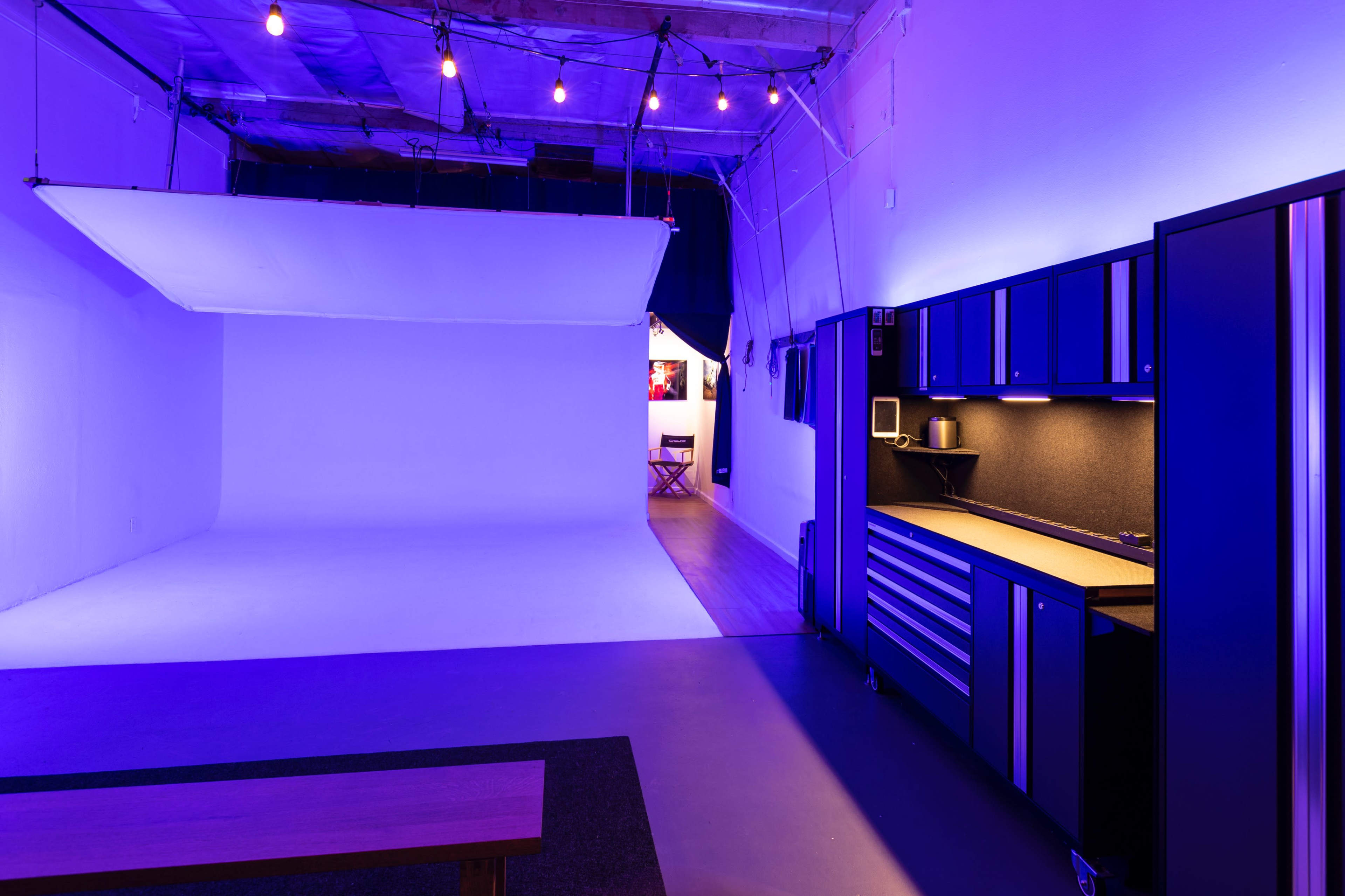 A photography studio with a white backdrop, a seating area, and a counter with storage cabinets, illuminated by soft purple lighting.