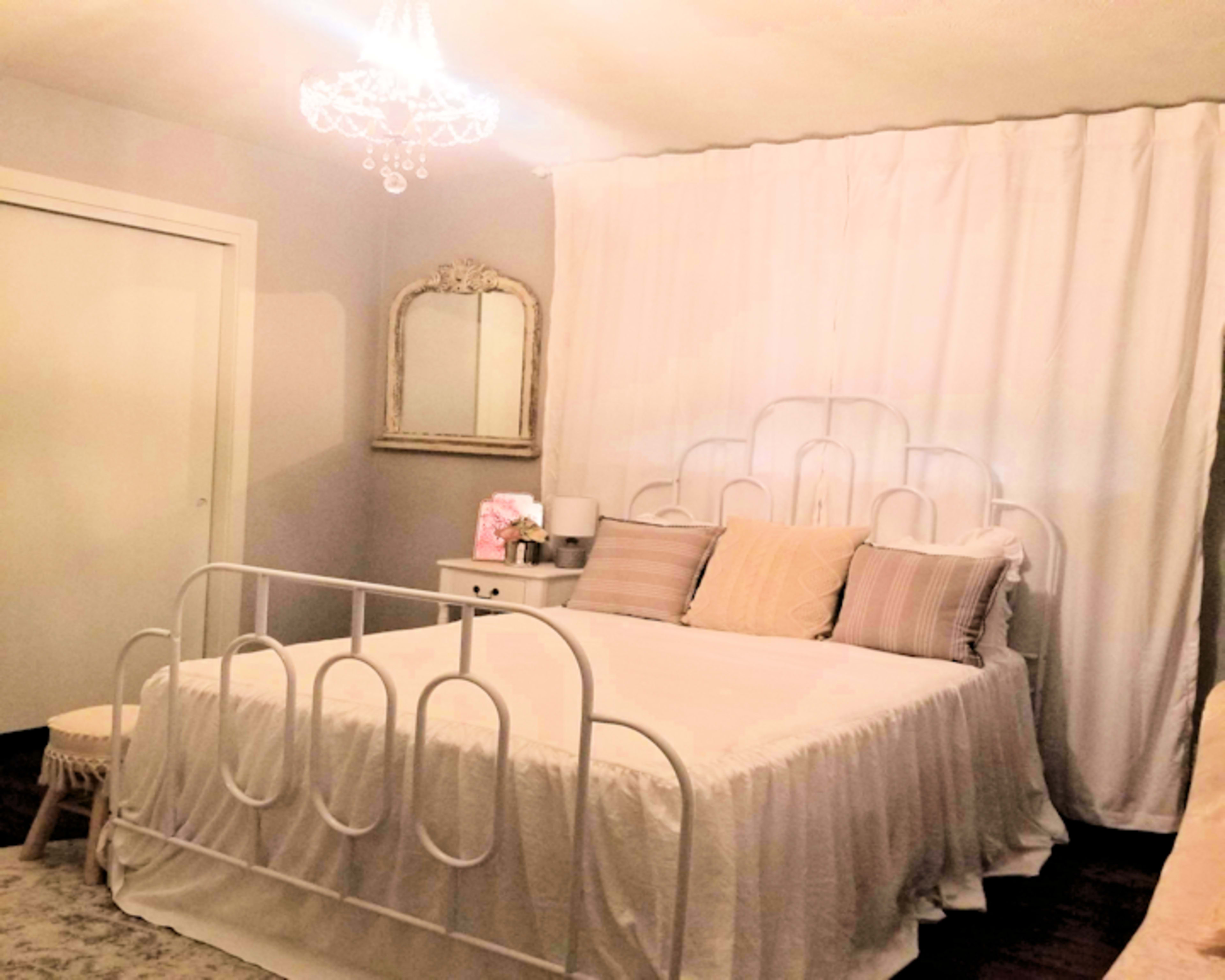 The room features a white bed with a decorative headboard, a chandelier overhead, and light-colored curtains hanging behind it.
