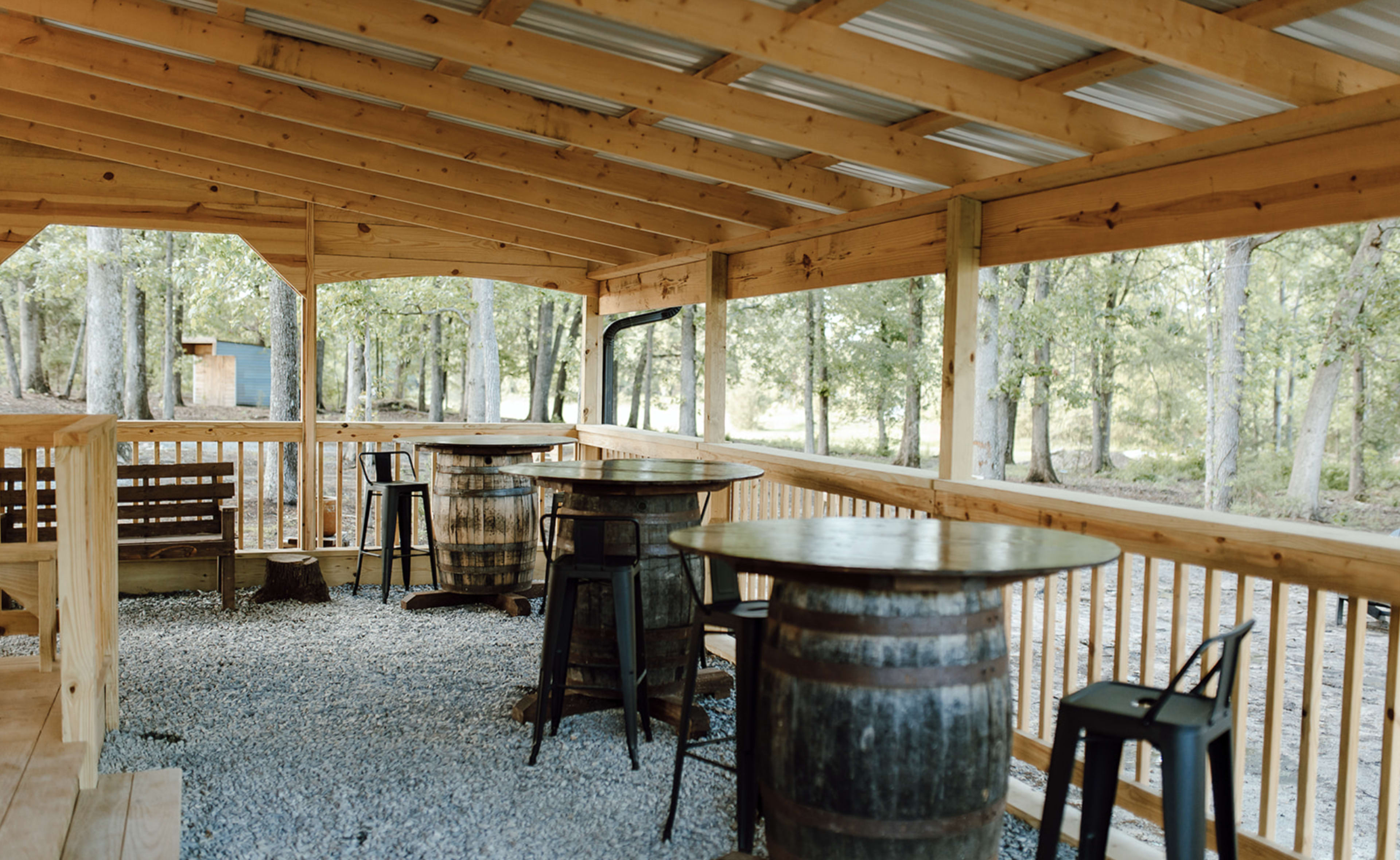 The image features a wooden gazebo-style structure with several tables and chairs set up on a gravel floor, surrounded by trees.