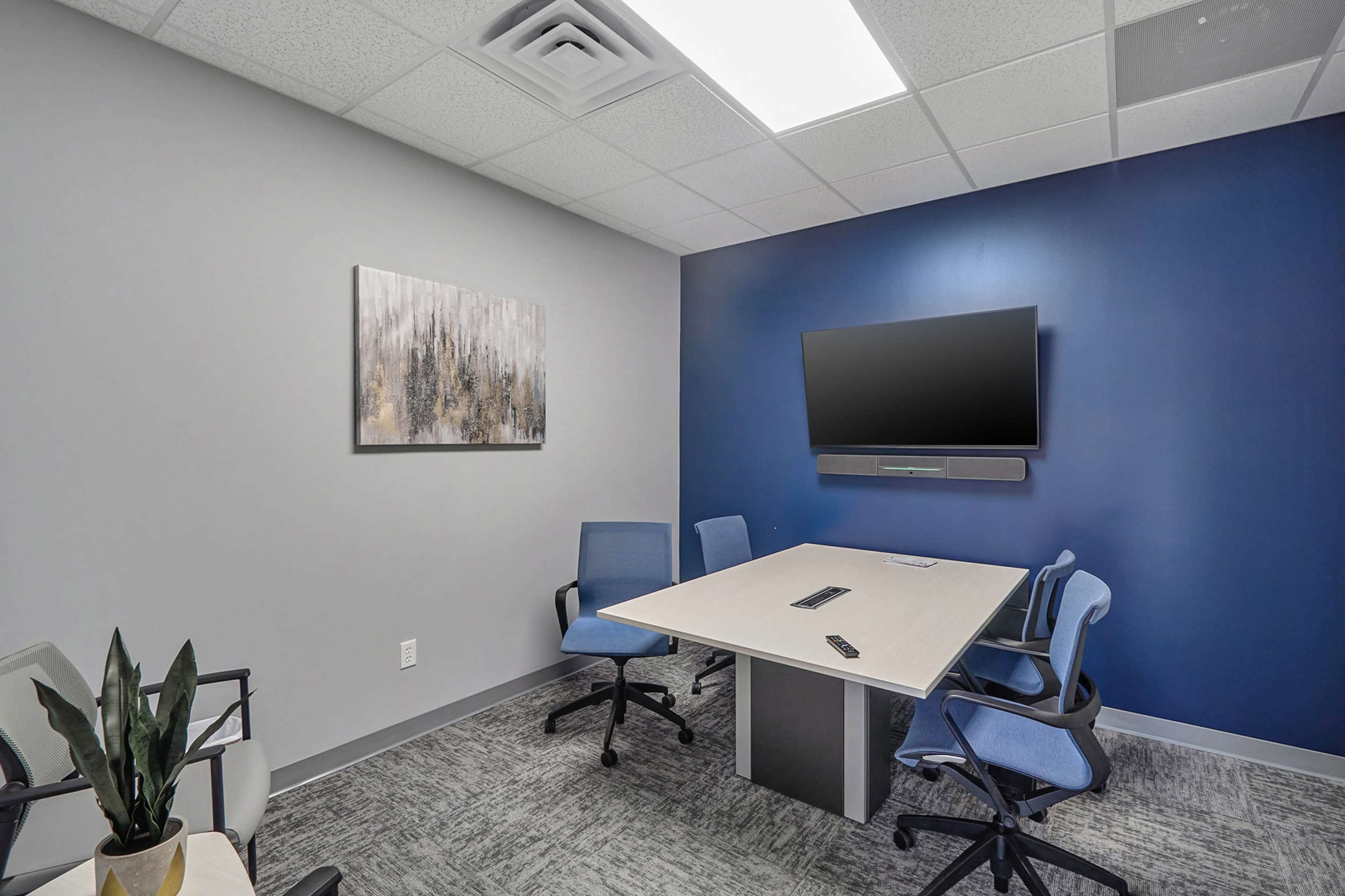 A modern conference room features a table surrounded by blue chairs, a wall-mounted television, and abstract artwork on a gray wall.
