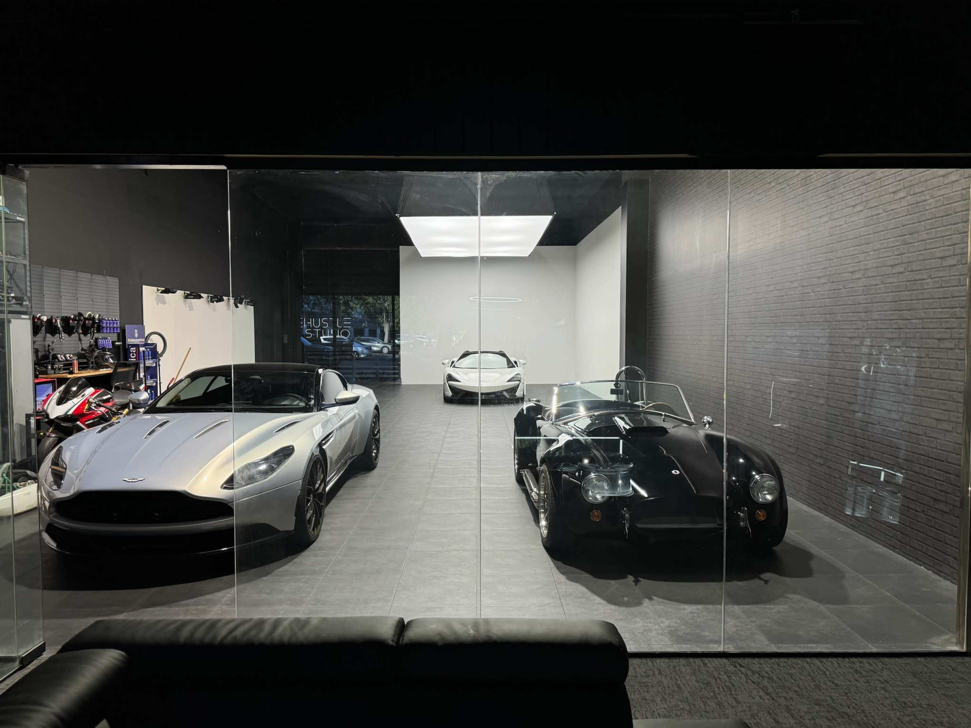 The image shows three luxury cars displayed in a glass-fronted showroom, with one convertible sports car on the left, a sleek coupe in the center, and a classic racing car on the right.