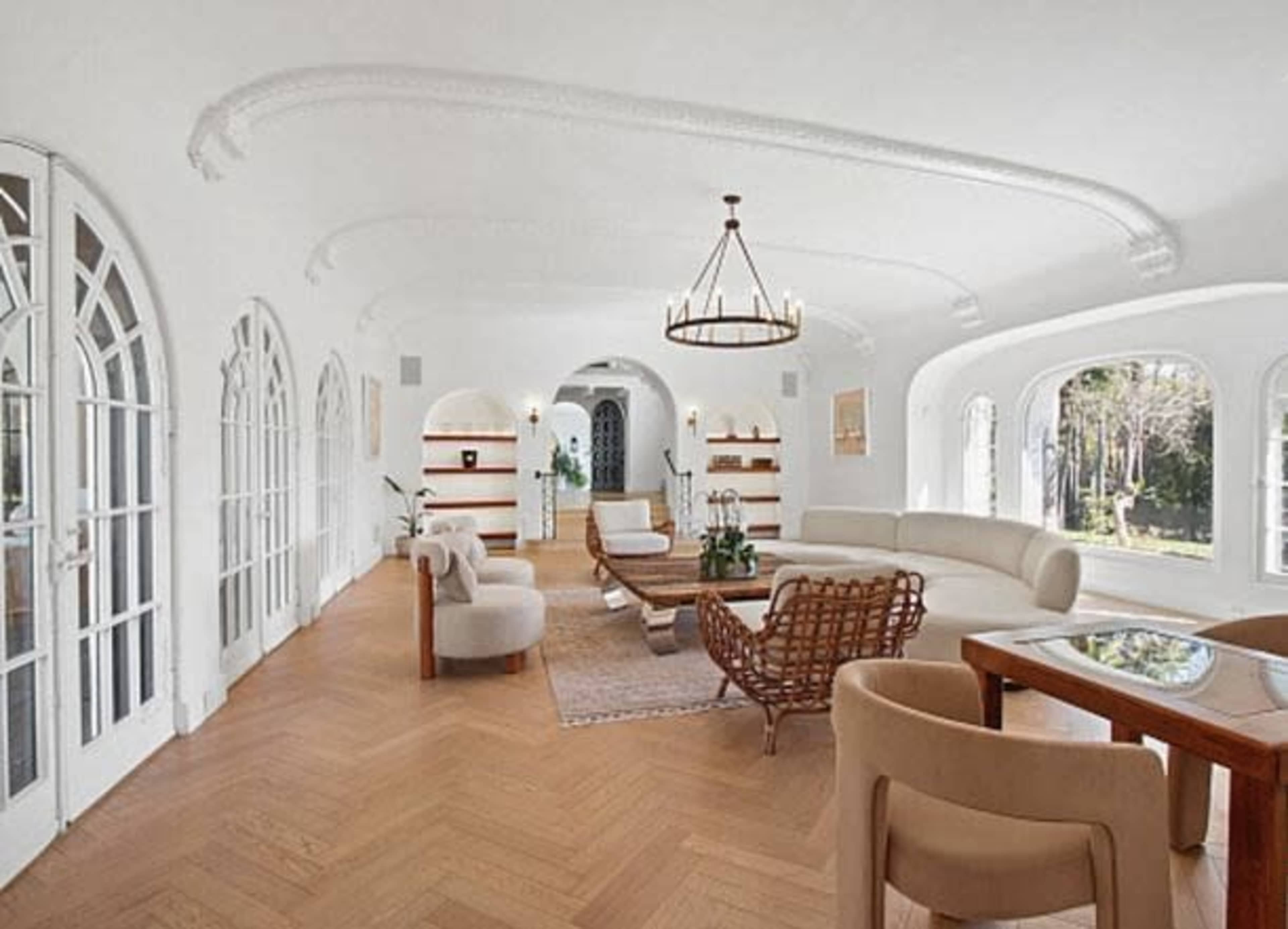 The image shows a spacious, elegantly designed living room with large windows, light-colored walls, and hardwood floors, featuring a mix of modern and traditional furniture.