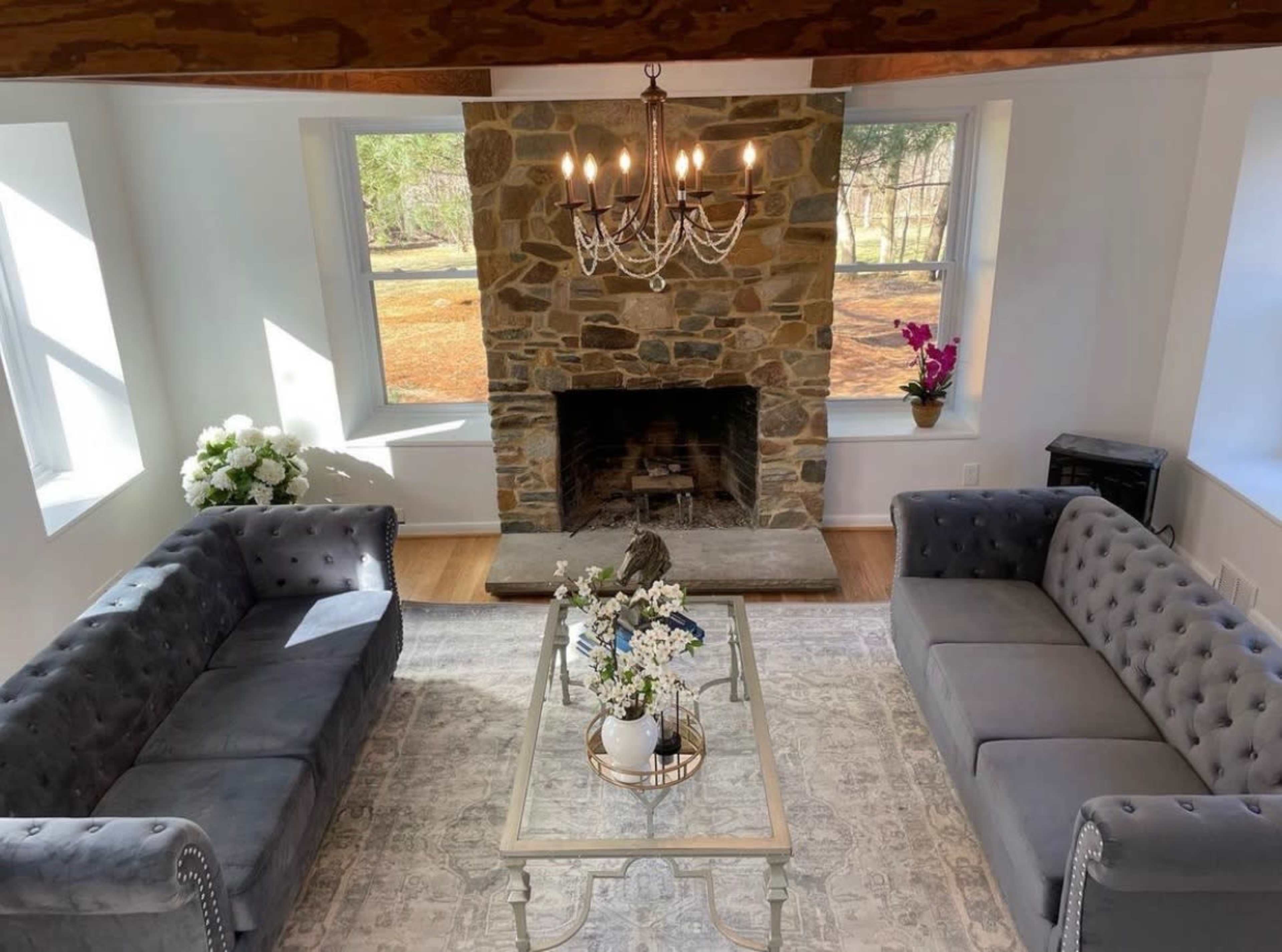 A bright living room featuring two gray tufted sofas, a coffee table with floral arrangements, and a stone fireplace beneath a chandelier.