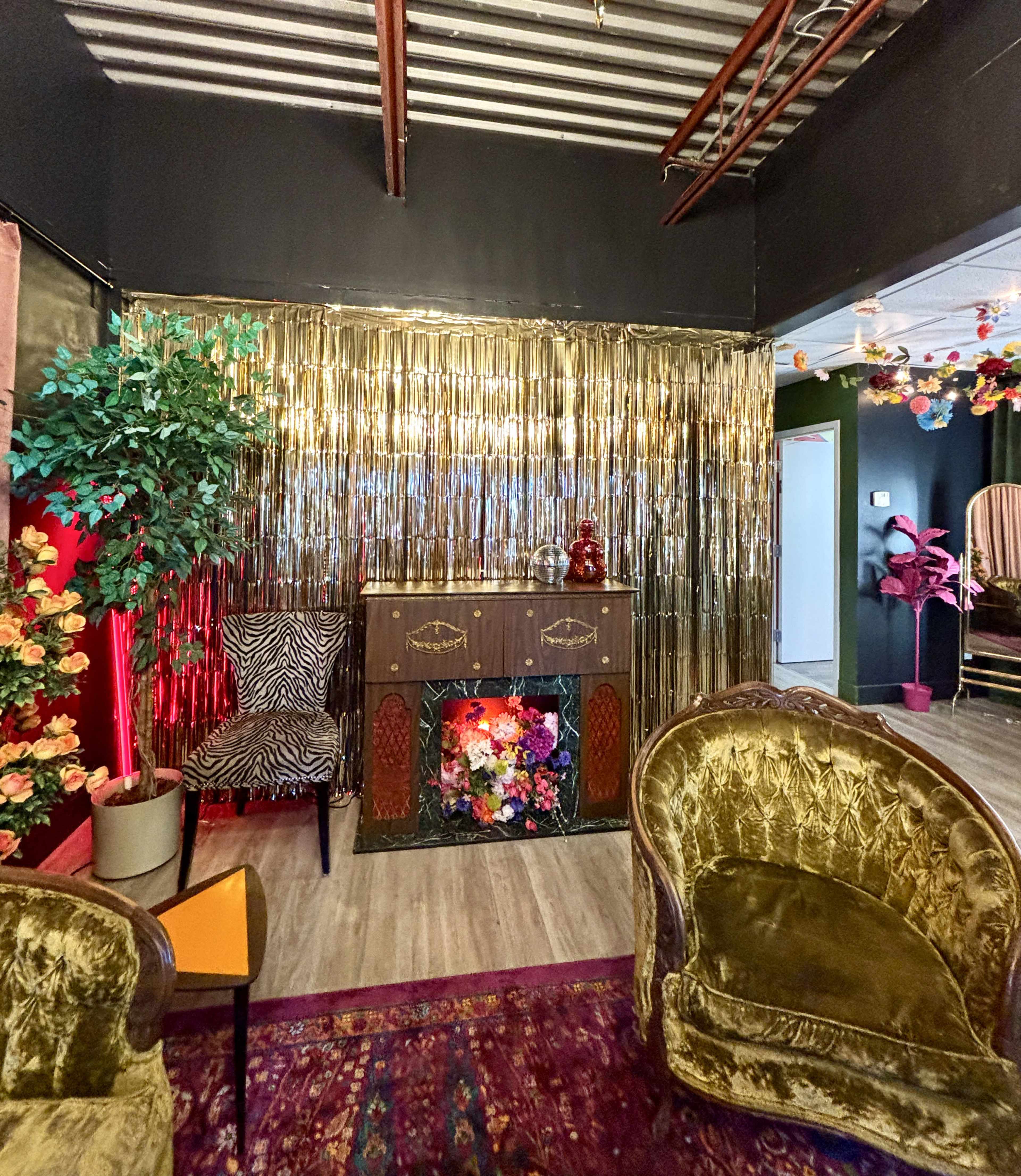 The image shows a vibrant interior of a styled room with a gold fringe backdrop, decorative chairs, and a floral arrangement on a small cabinet.