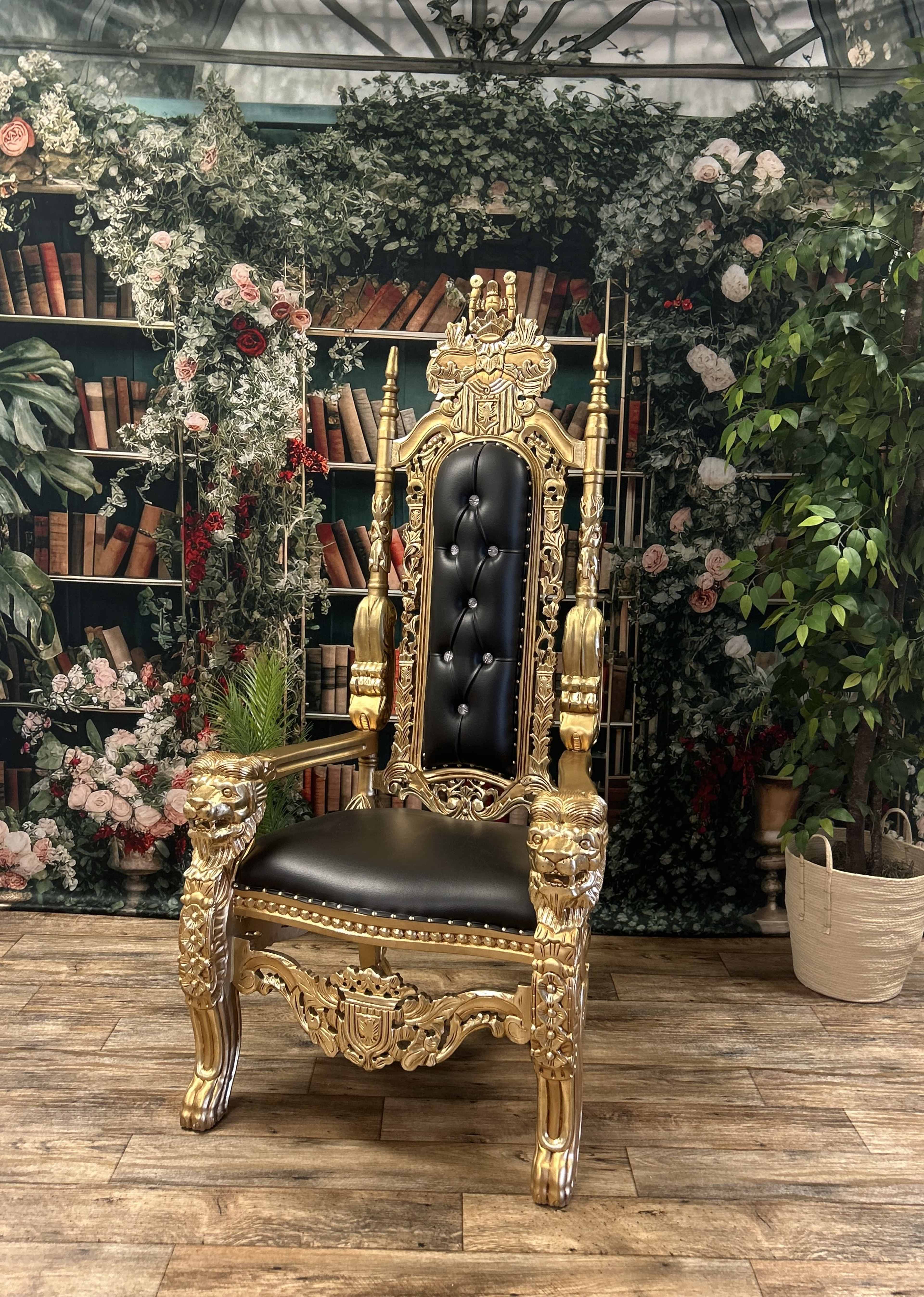 The image depicts an ornate golden throne with a black upholstered seat, placed against a backdrop of shelves filled with books and greenery.