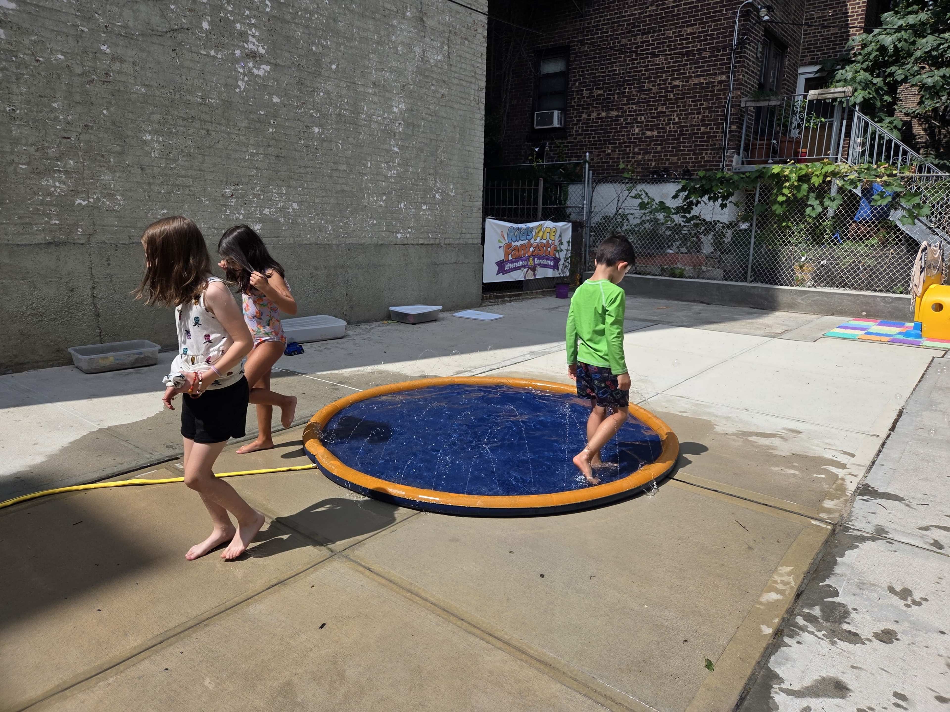 Three children play around a circular inflatable water pool in a concrete yard, with a brick wall and playground equipment in the background.