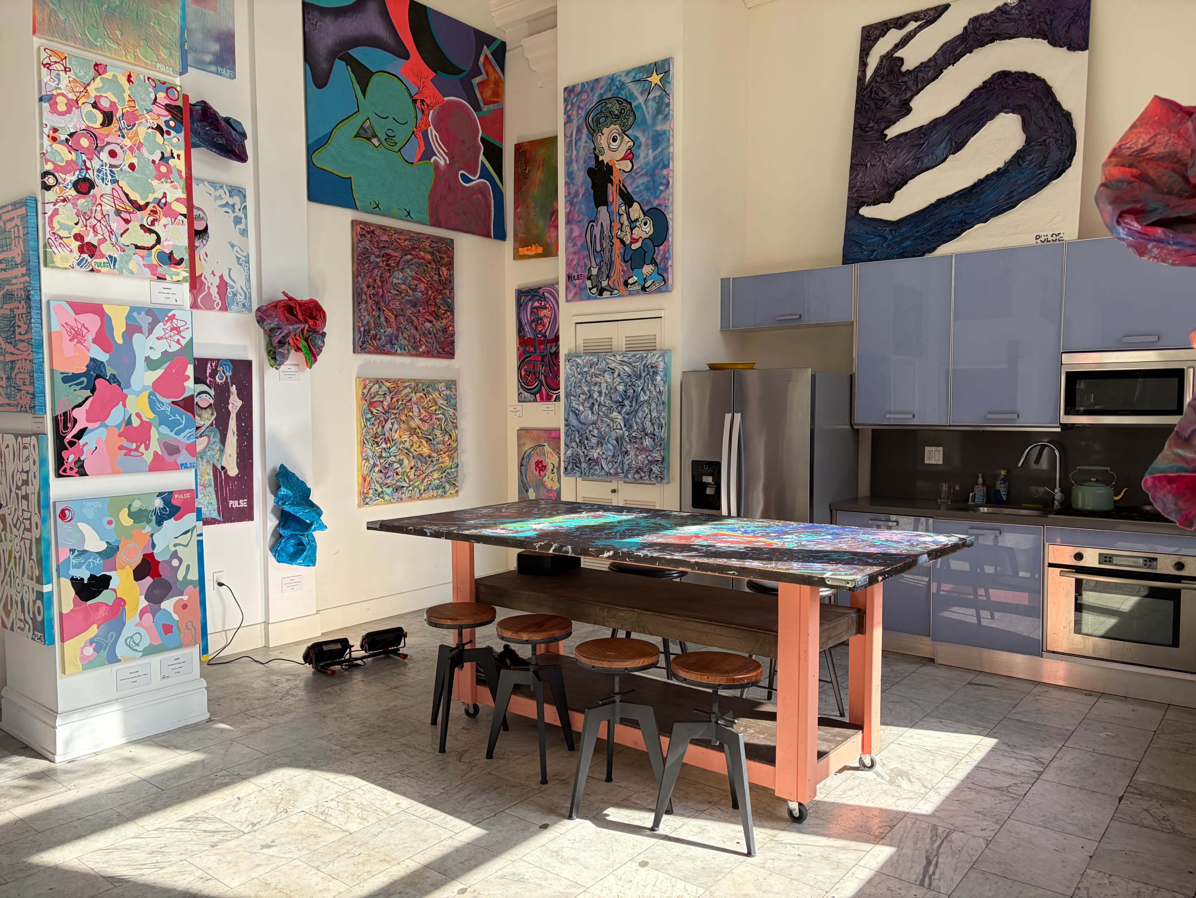 The image shows an art studio featuring colorful paintings on the walls, a modern kitchen area with appliances, and a large central table surrounded by stools.