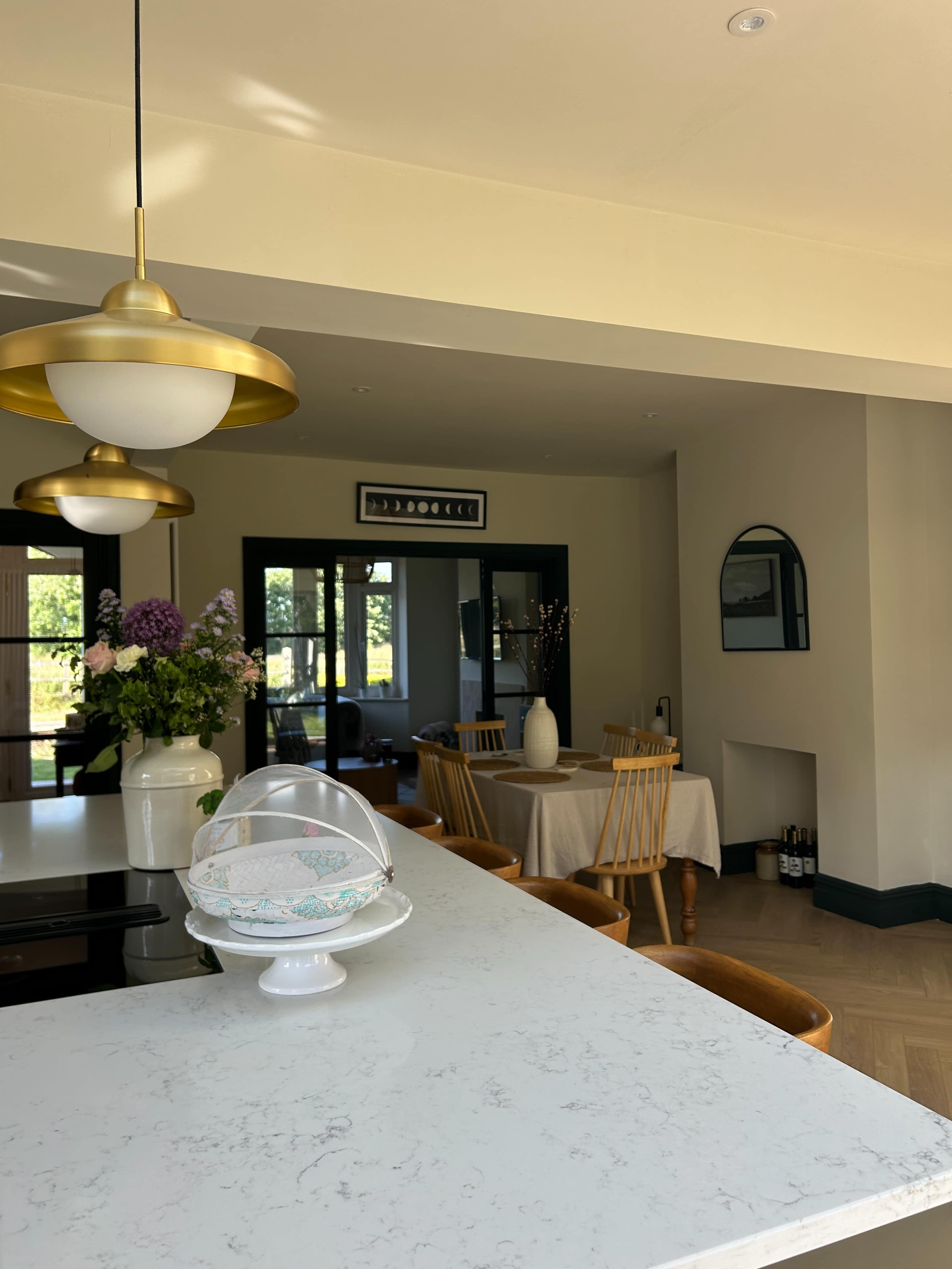 The image shows a bright kitchen and dining area featuring a marble countertop, gold light fixtures, and a table set for dining with wooden chairs.